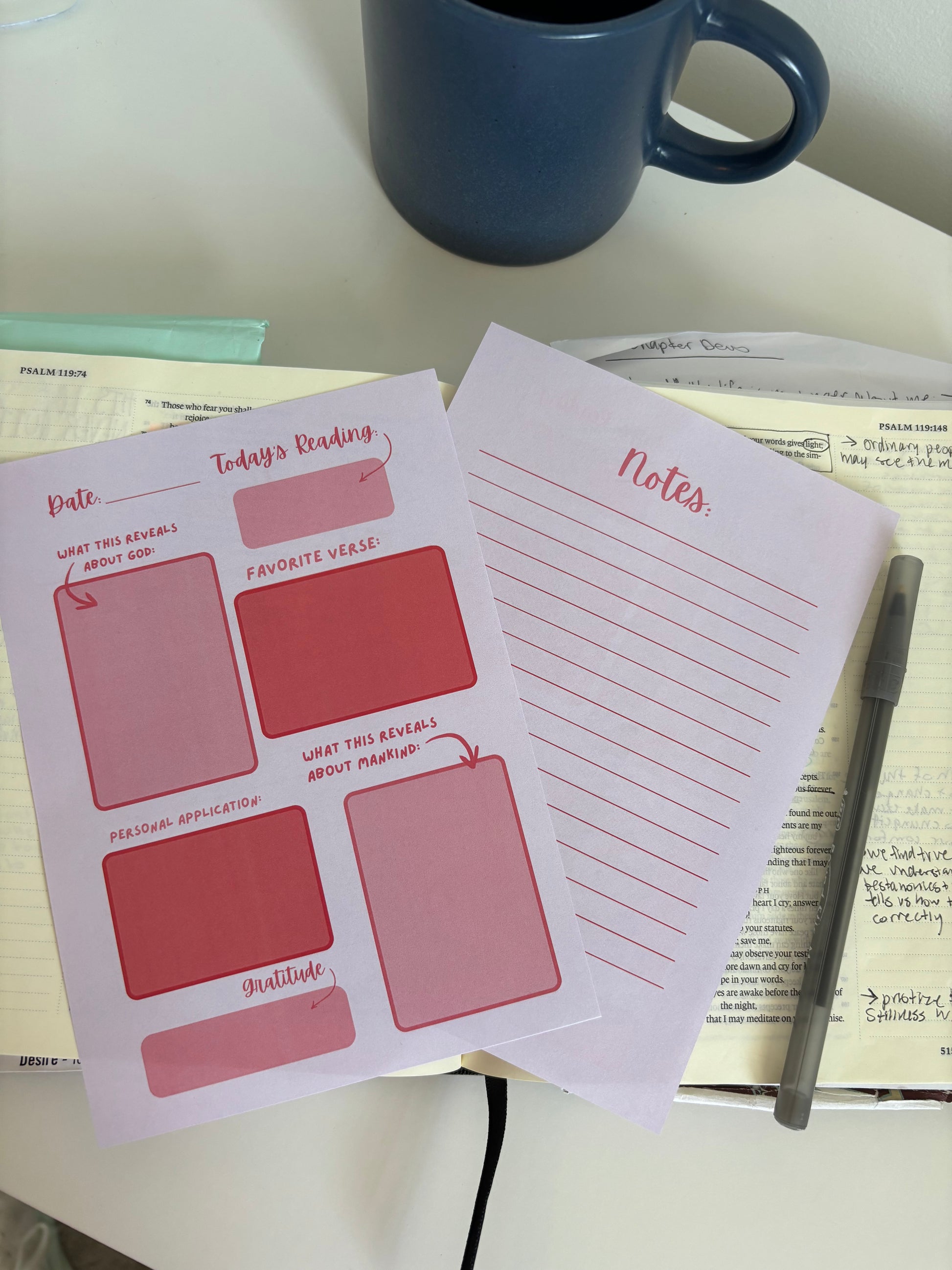 Notebook pages with red and pink design on a desk with a blue mug and pen.