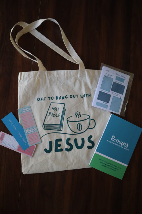 Tote bag with 'Jesus' text and graphic, surrounded by small books or cards on a wooden surface.