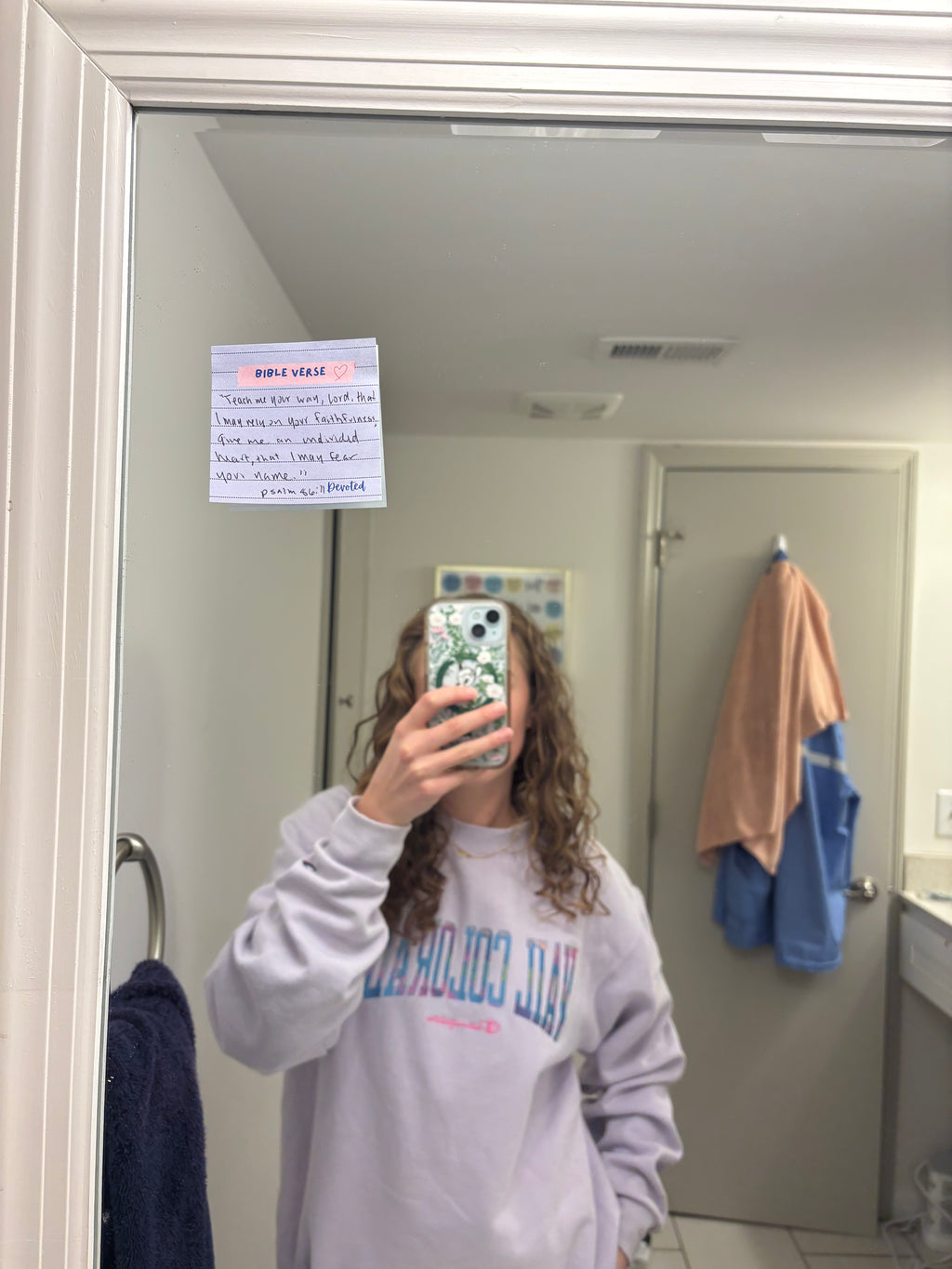 Person taking a mirror selfie wearing a sweatshirt with text, in a bathroom setting.