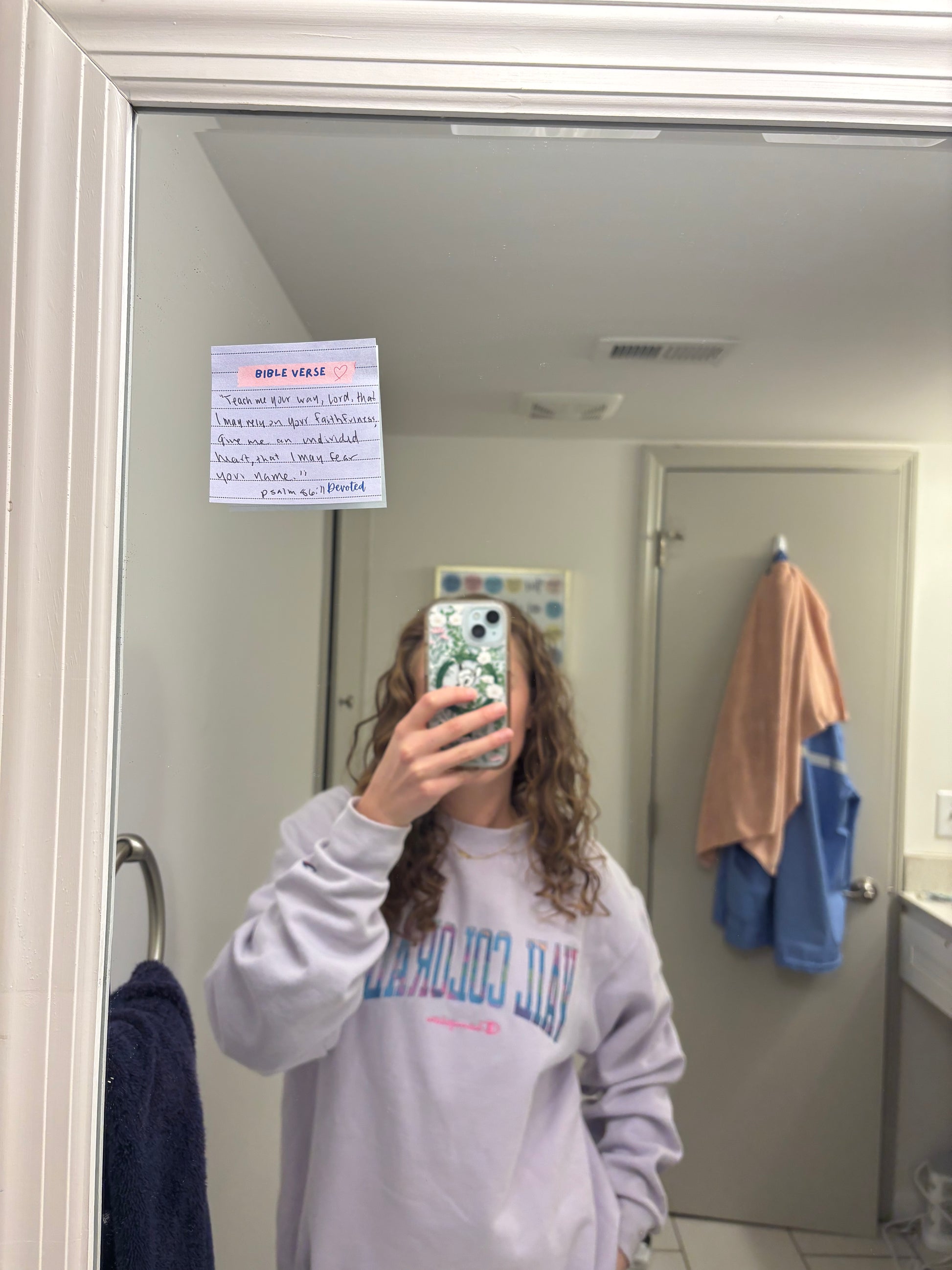 Person taking a mirror selfie wearing a sweatshirt with text, in a bathroom setting.