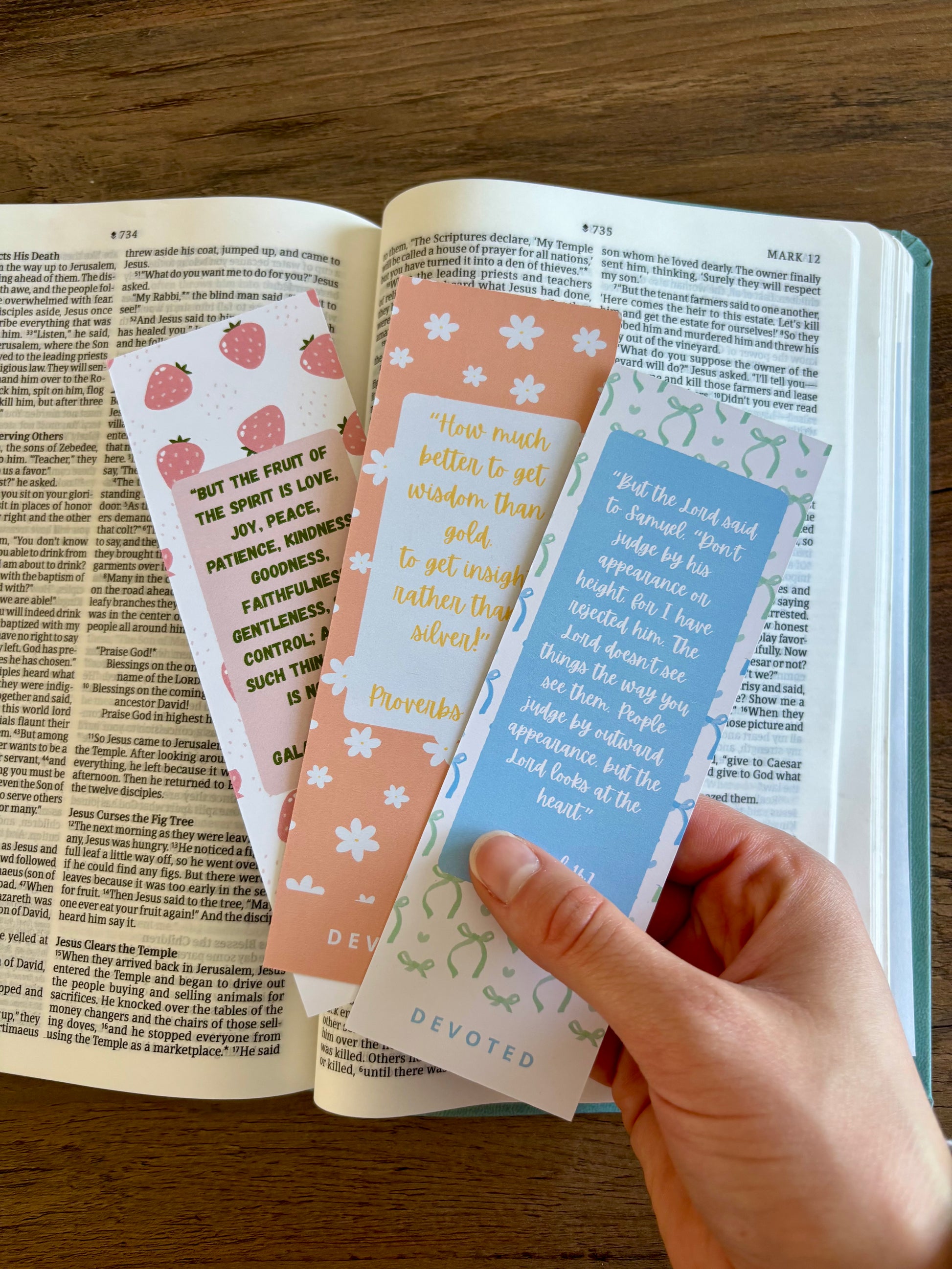 Hand holding three colorful bookmarks in front of an open book on a wooden surface