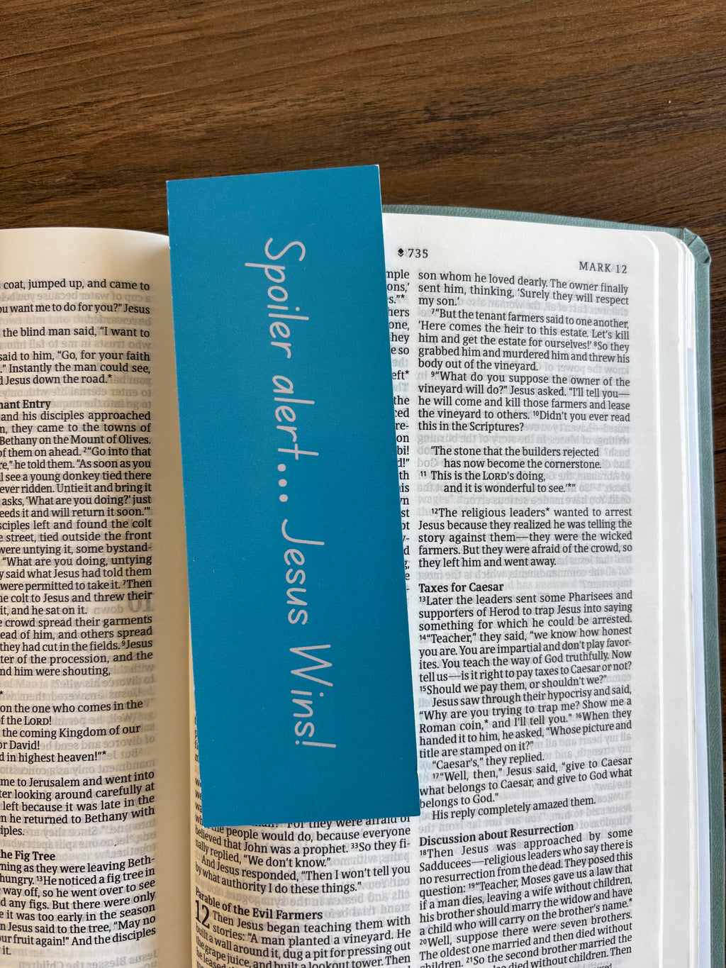 Open book with a blue bookmark titled 'Spoiler alert... Jesus Wins!' on a wooden surface