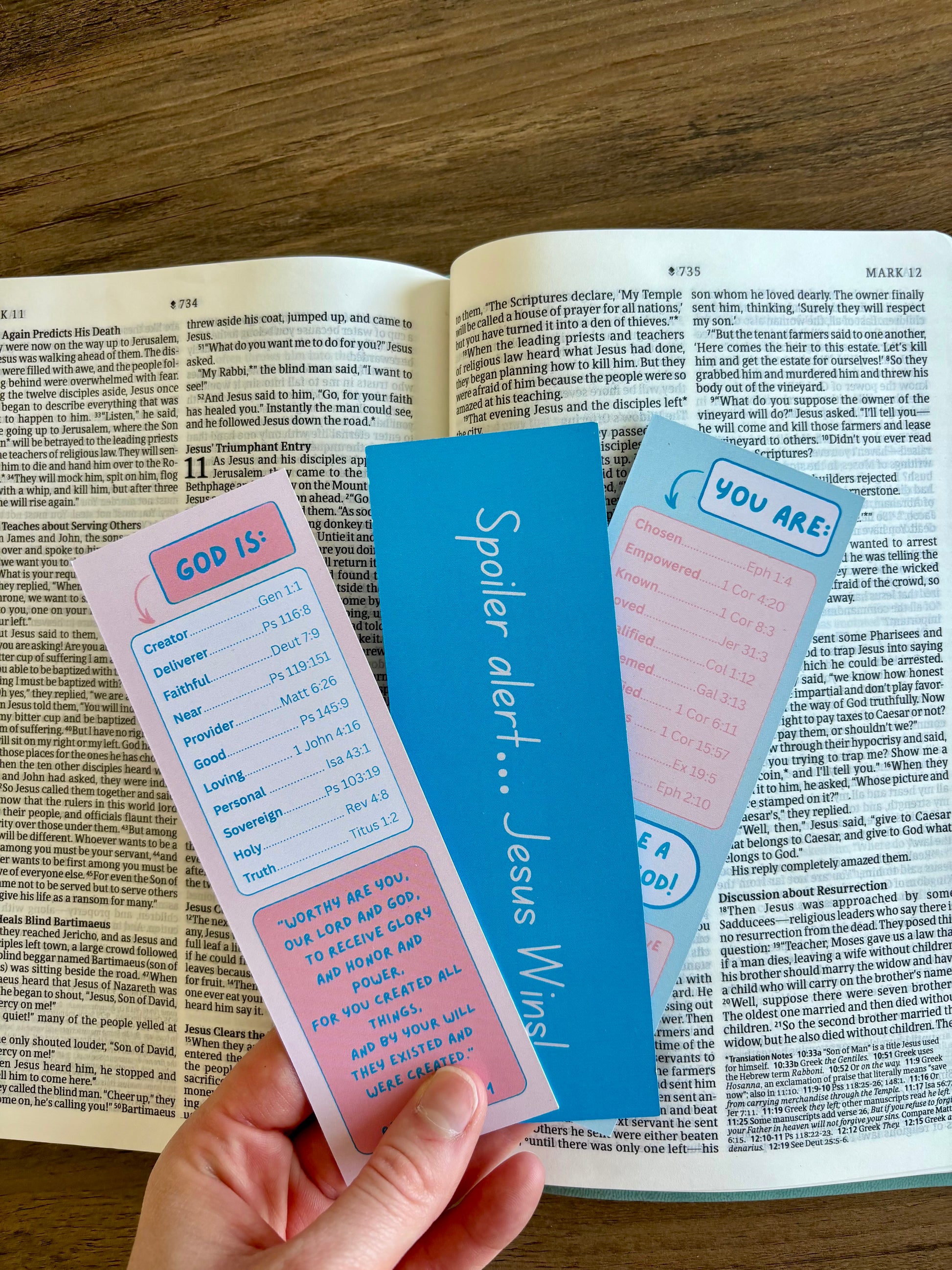 Three bookmarks with text held over an open book on a wooden surface