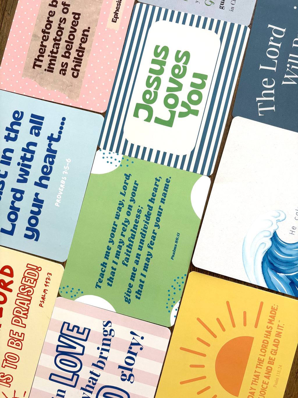 A close-up, angled view of multiple colorful Bible verse cards arranged on a dark wooden surface. The cards feature various designs including stripes, polka dots, sun graphics, and text in blue, green, pink, yellow, and white.