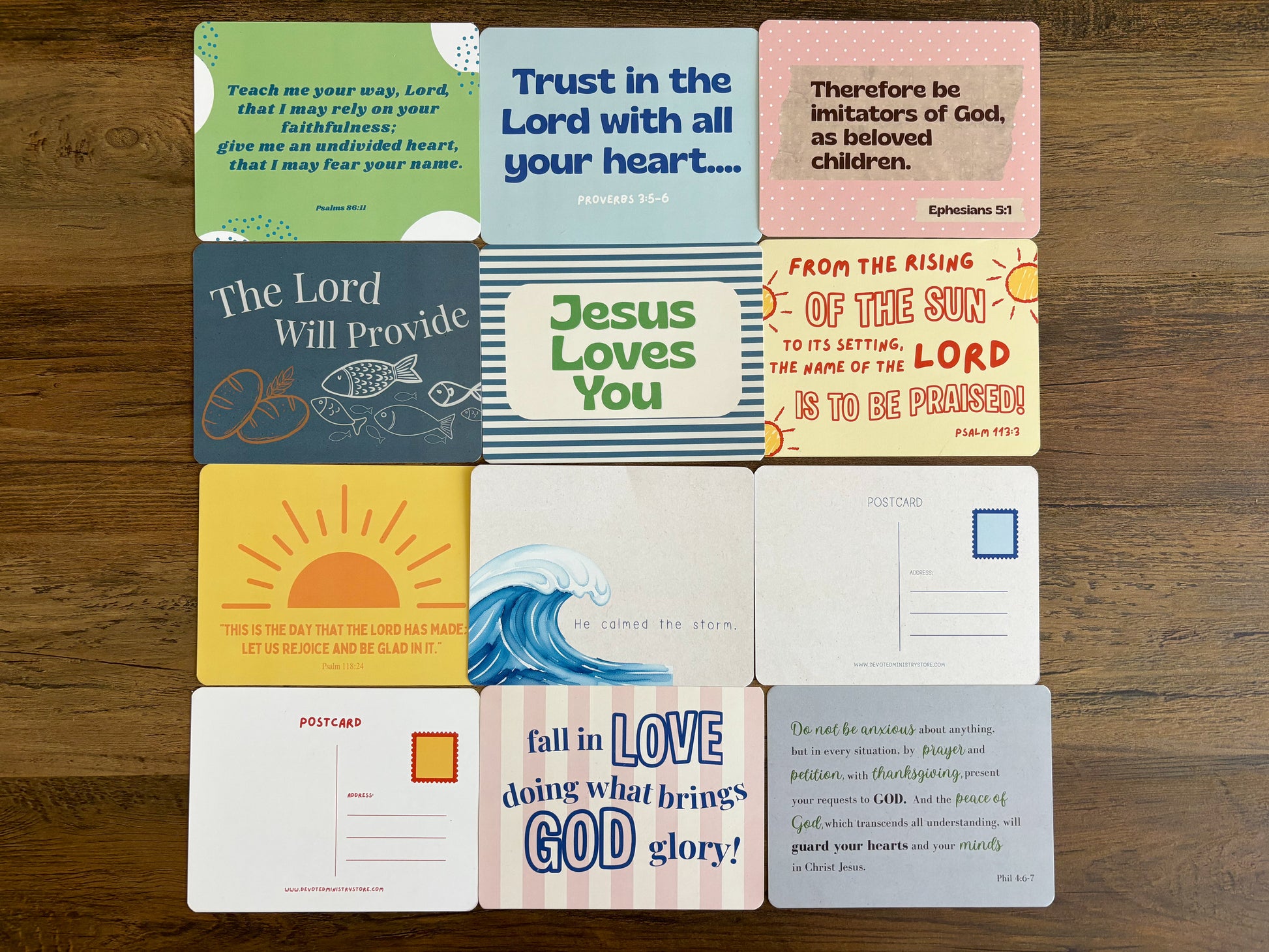 A 3x3 grid of square postcards with various colorful Bible verses, displayed flat on a dark wood background.