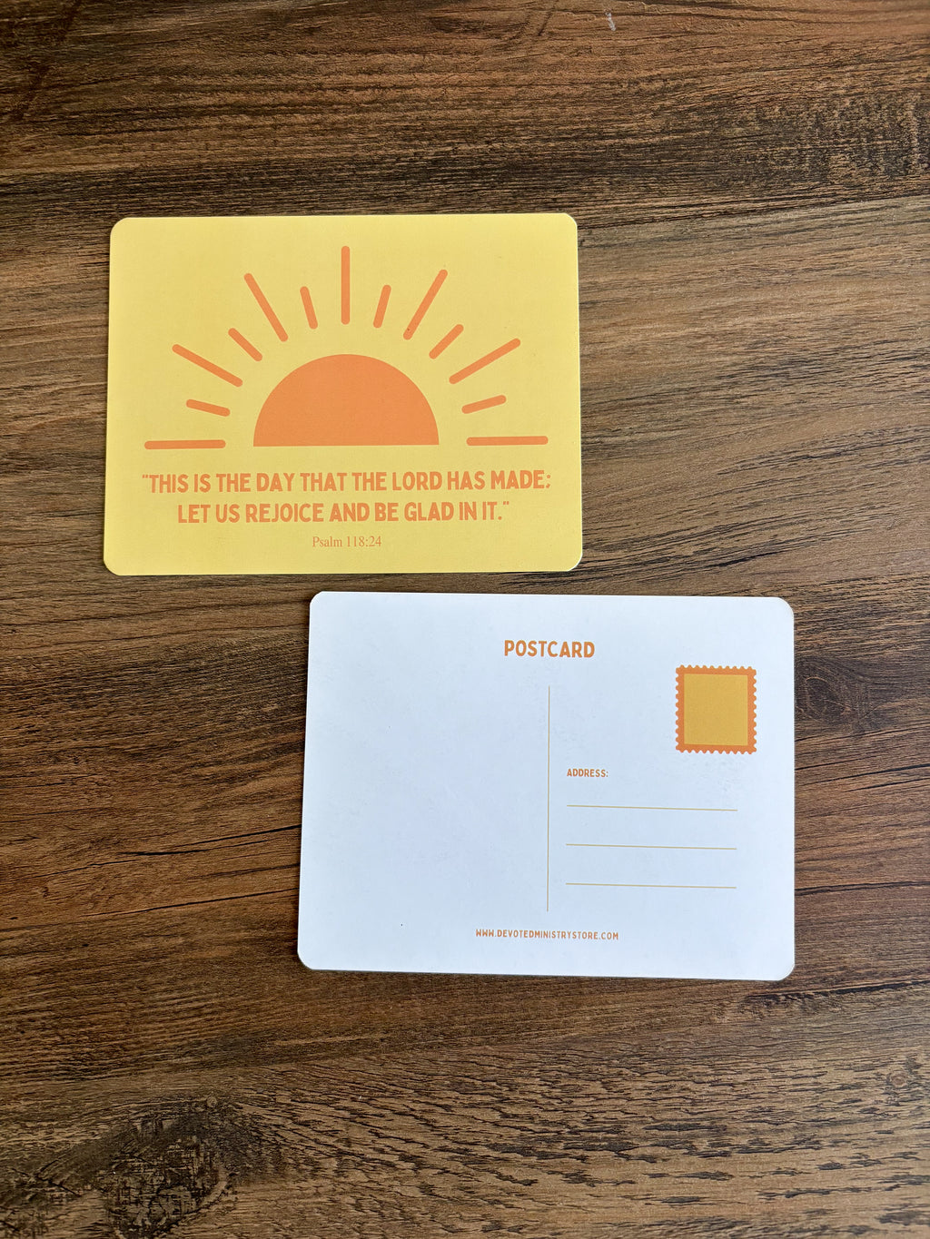 A yellow postcard with an orange sun graphic and the Bible verse Psalm 100:2 printed on the front, shown above a white postcard back on a dark wood surface.