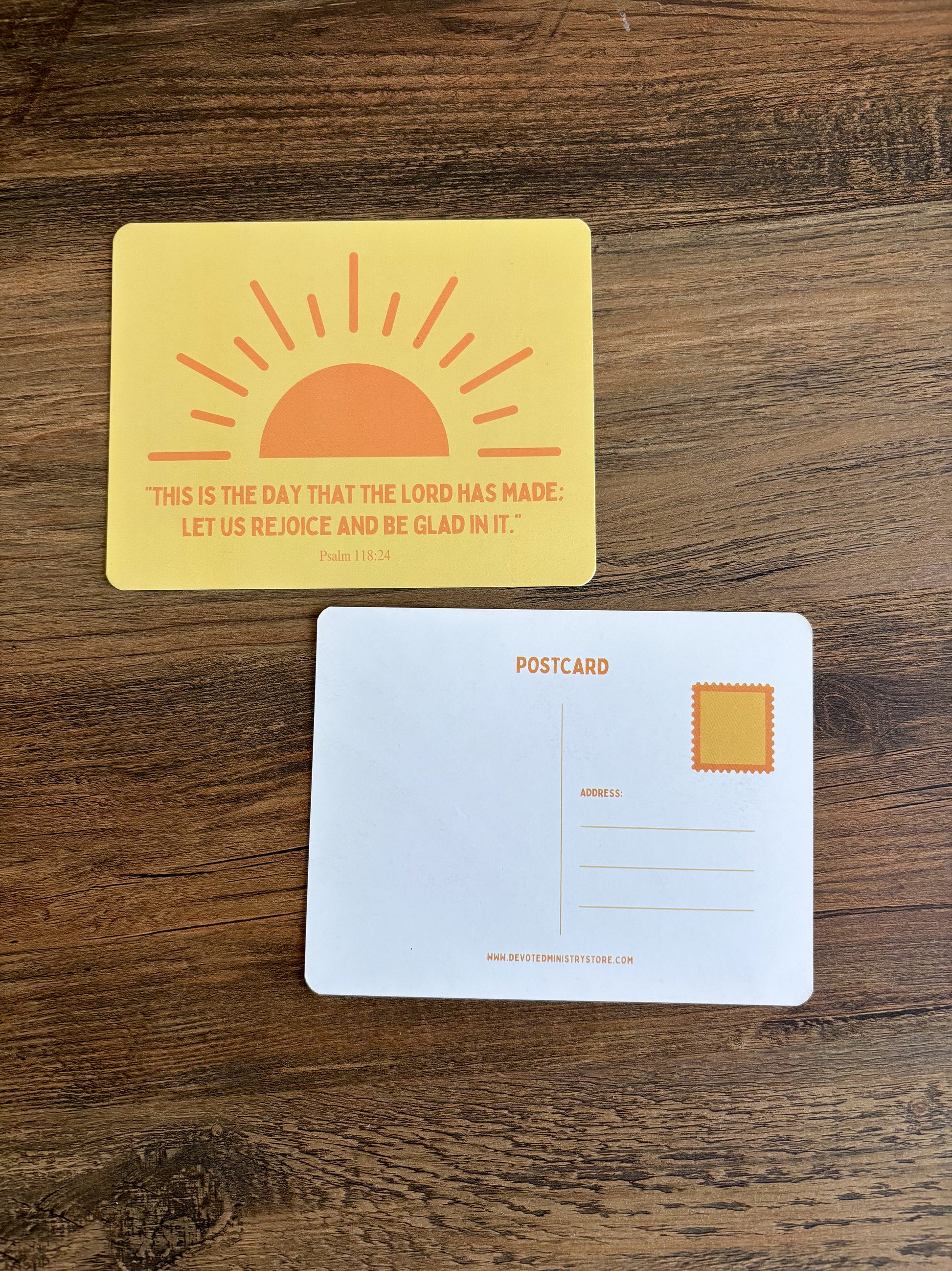 A yellow postcard with an orange sun graphic and the Bible verse Psalm 100:2 printed on the front, shown above a white postcard back on a dark wood surface.