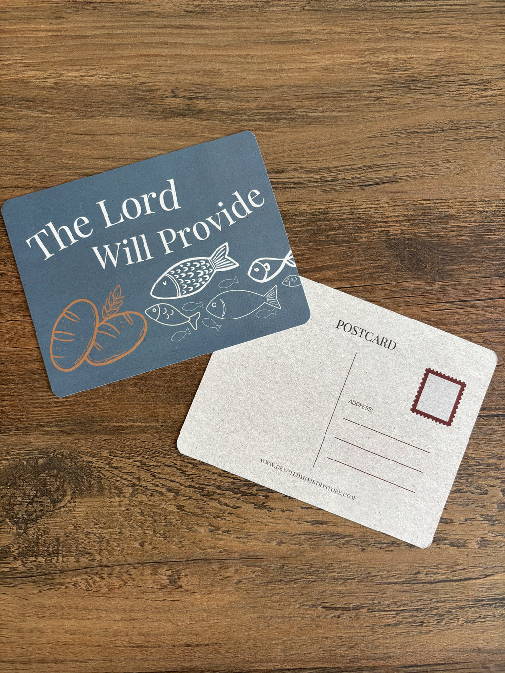 A postcard-style gift set on a wooden tabletop, featuring one blue front card with the text 'The Lord Will Provide' and simple food illustrations, and one white back card with postcard lines and a red stamp box.