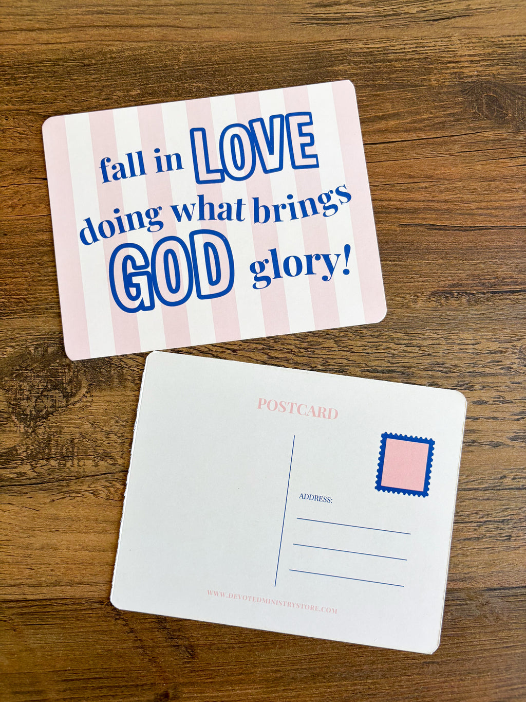 Two postcards displayed on a wooden surface. The top postcard has pink and white vertical stripes with blue text reading 'fall in LOVE doing what brings GOD glory!'.