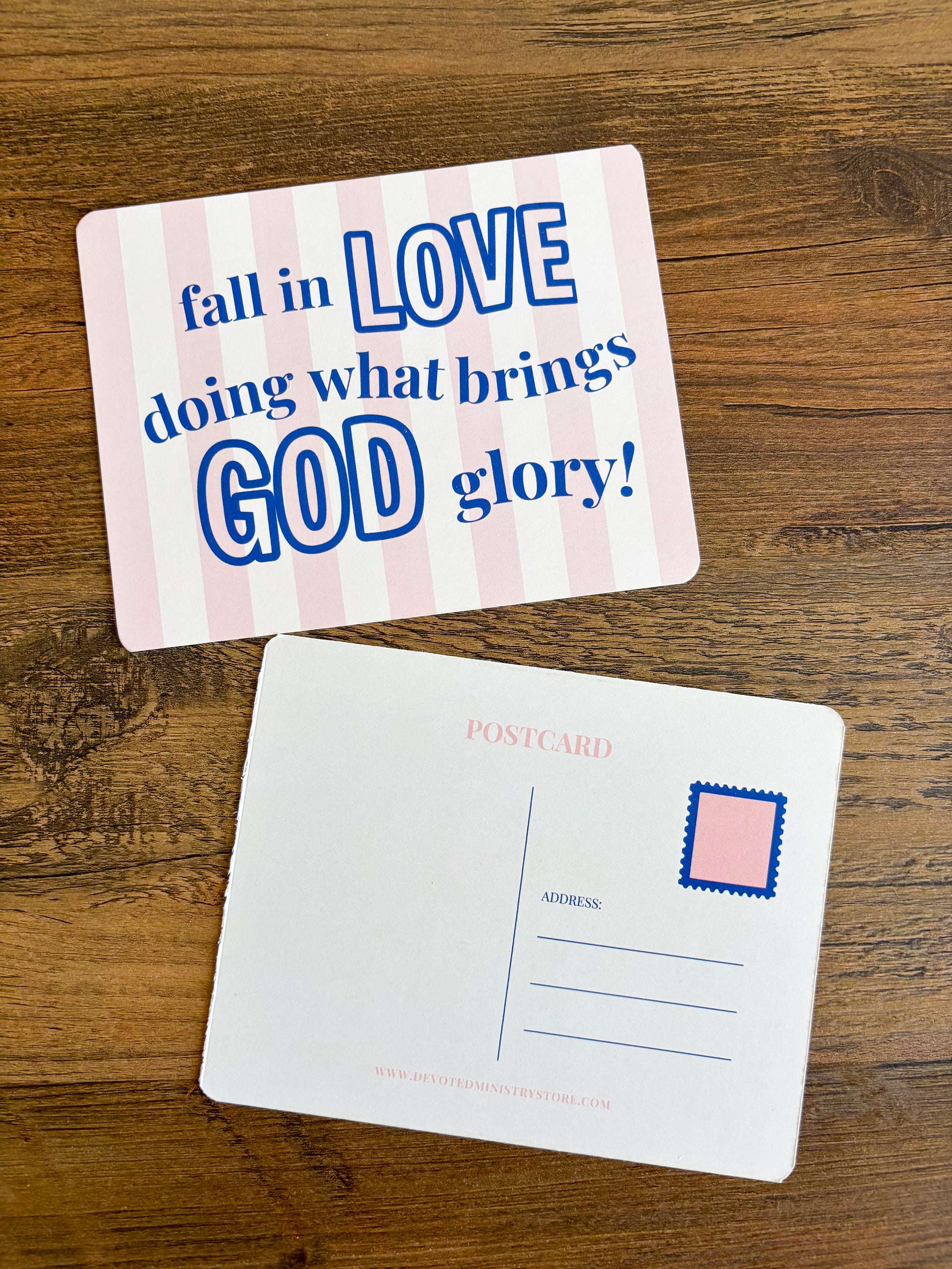 Two postcards displayed on a wooden surface. The top postcard has pink and white vertical stripes with blue text reading 'fall in LOVE doing what brings GOD glory!'.