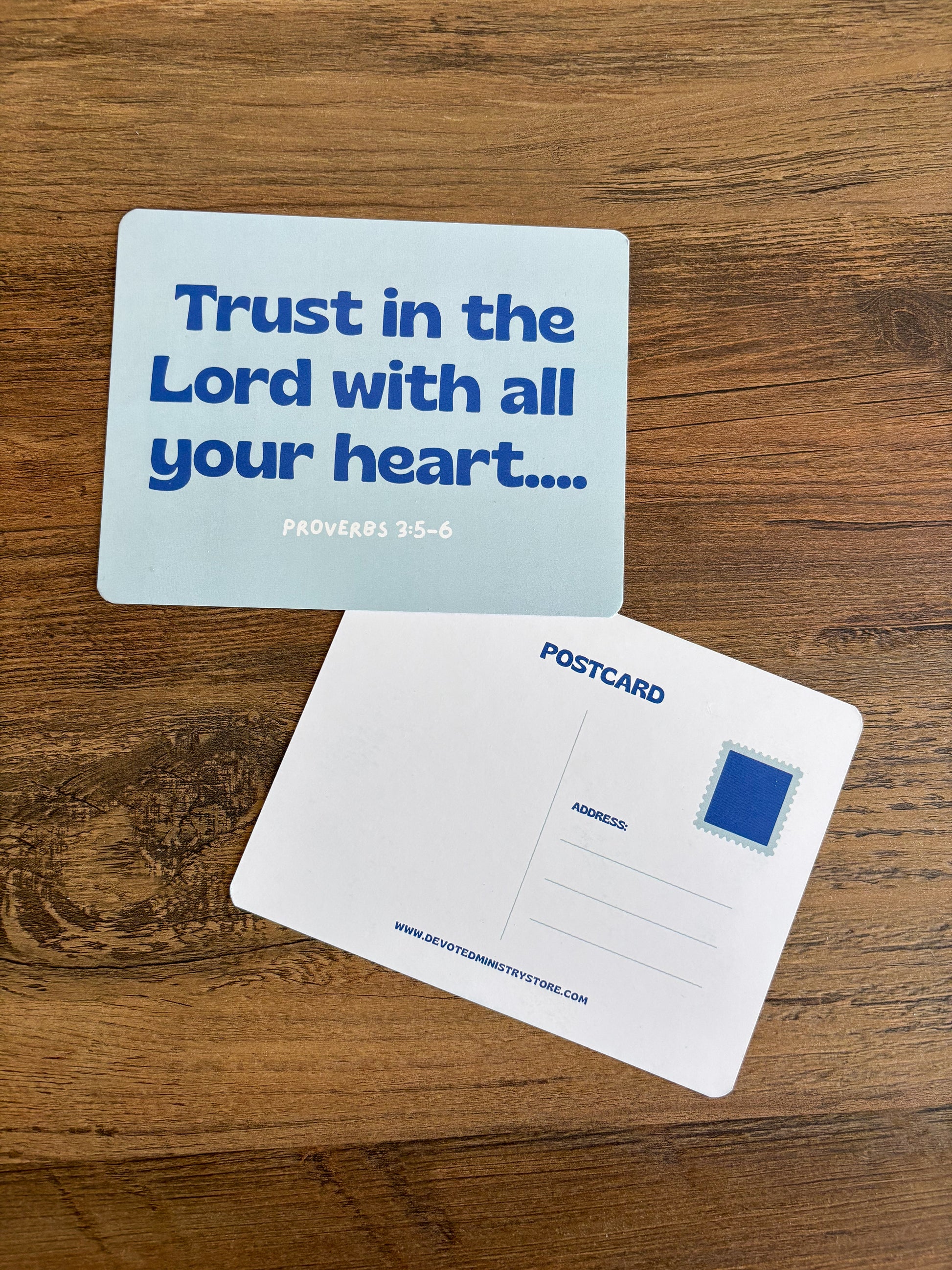 A postcard set on a wooden tabletop, showing the front with the blue text 'Trust in the Lord with all your heart.