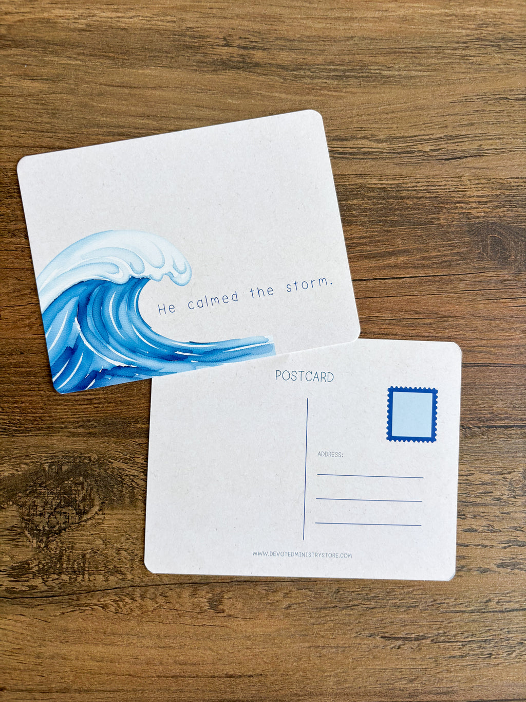 A postcard set displayed on a wooden surface, showing both the front and back. The front features a blue ocean wave illustration with the text 'He calmed the storm.