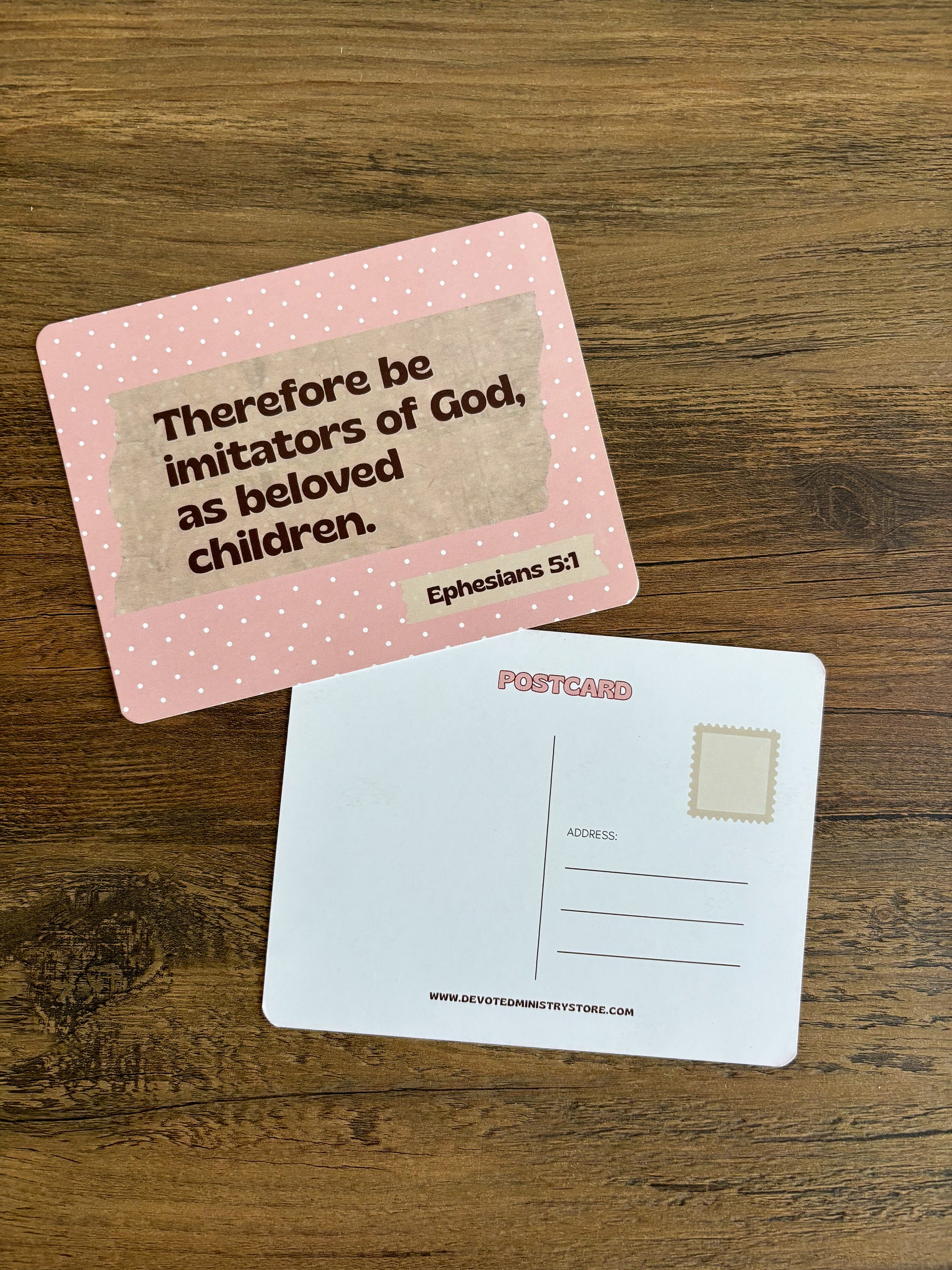 A pink postcard with a Bible verse from Ephesians 5:1 printed on the front, placed on top of a white postcard showing the back with address lines and a stamp box, all on a wooden surface.