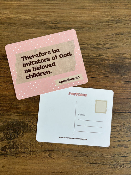 A pink postcard with a Bible verse from Ephesians 5:1 printed on the front, placed on top of a white postcard showing the back with address lines and a stamp box, all on a wooden surface.