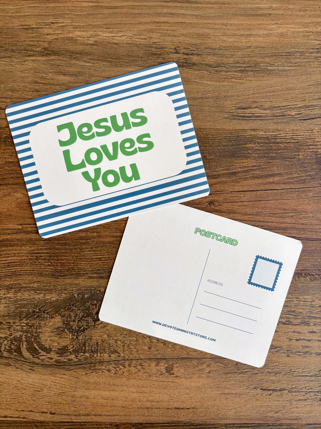 A postcard set on a wooden surface, showing the front with blue and white stripes and green text reading 'Jesus Loves You,' and the back with postcard fields and a blue stamp box.