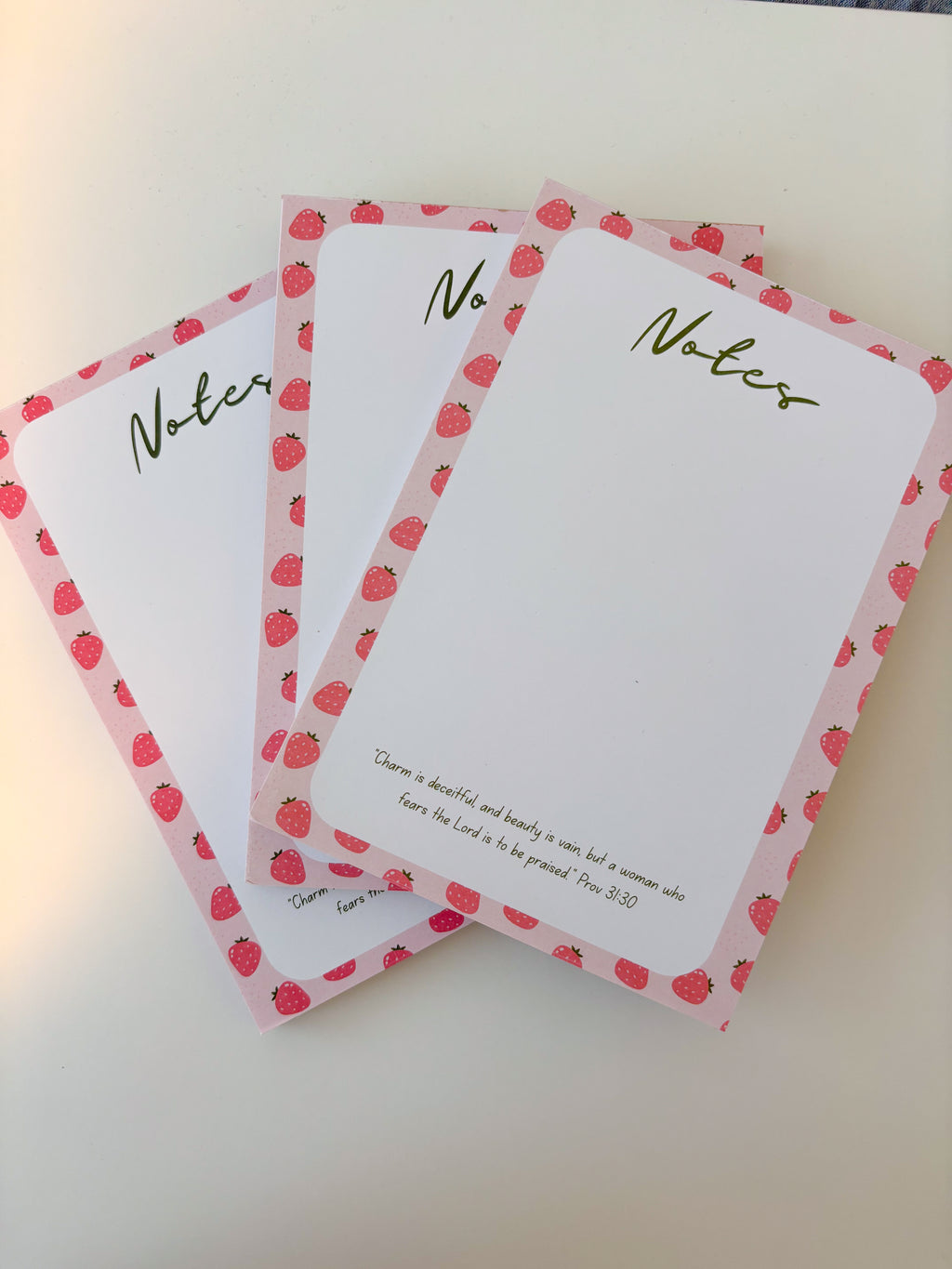 Three white notepad sheets with pink strawberry-patterned borders and green 'Notes' lettering, arranged on a light beige surface.