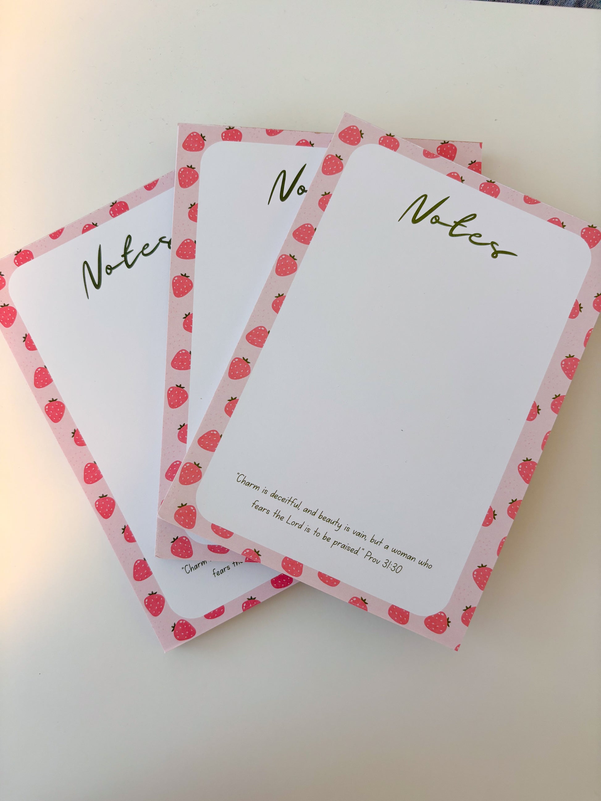 Three white notepad sheets with pink strawberry-patterned borders and green 'Notes' lettering, arranged on a light beige surface.