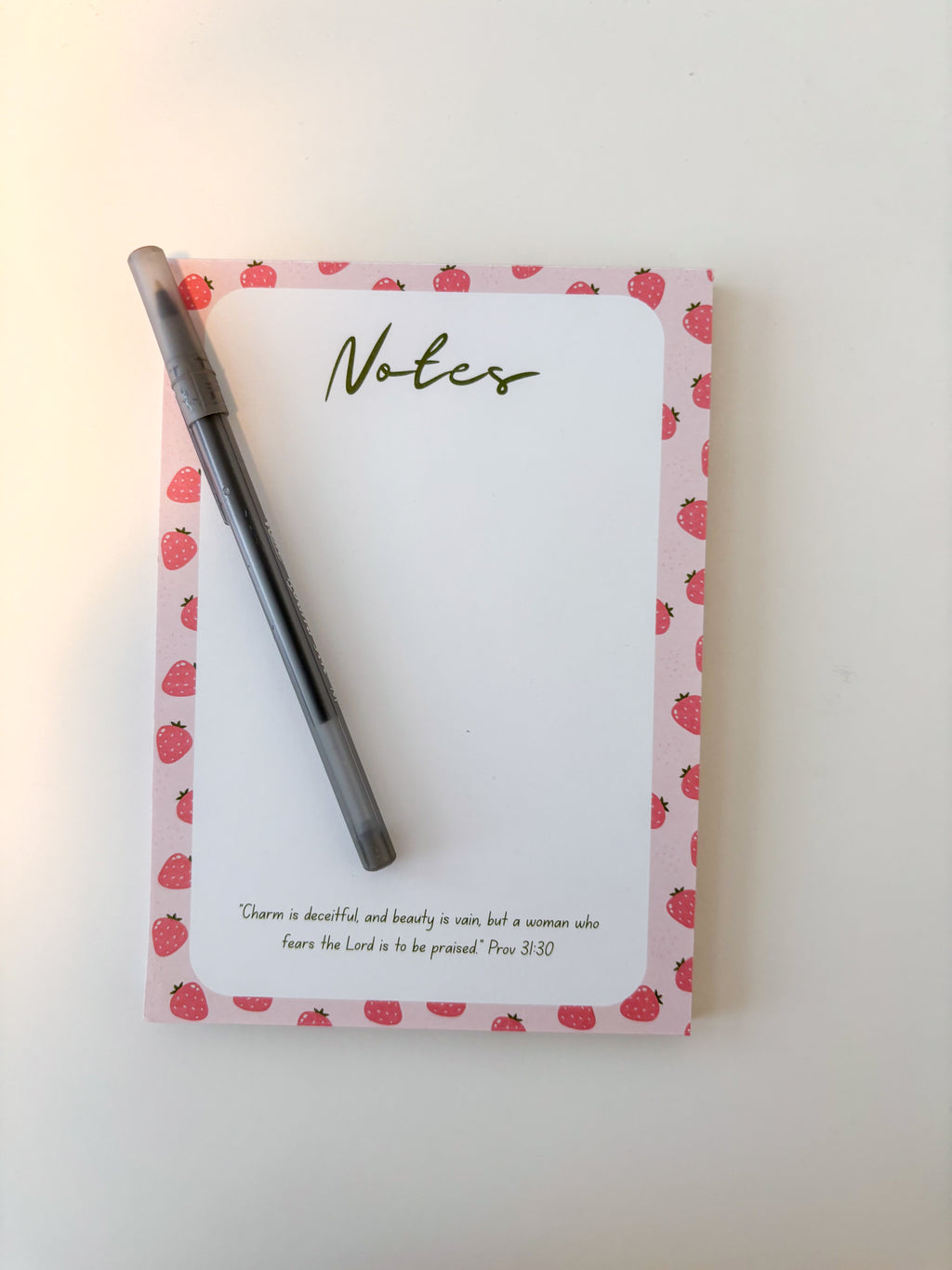 A pink notepad with a strawberry pattern border and a black pen placed on top, photographed on a plain light background.