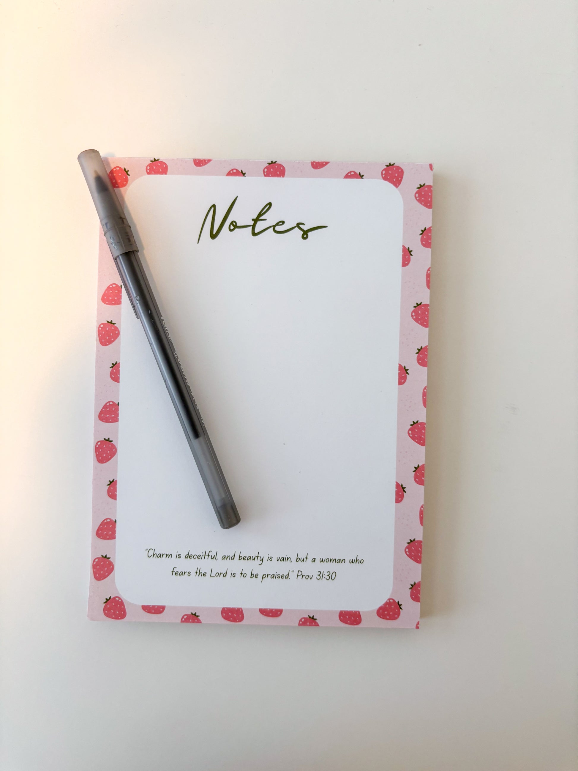 A pink notepad with a strawberry pattern border and a black pen placed on top, photographed on a plain light background.
