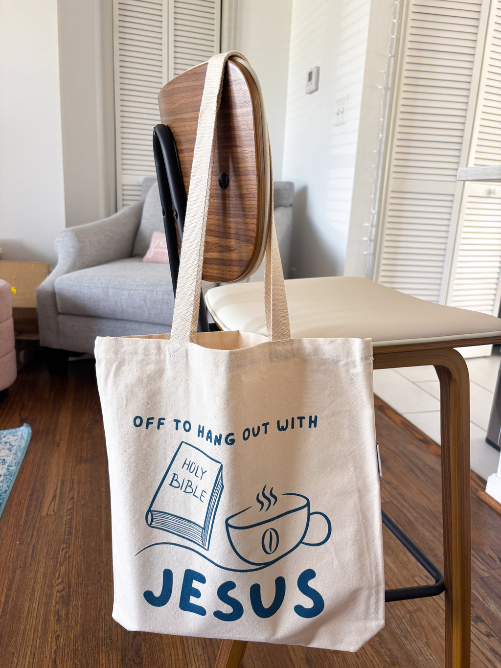 A cream canvas tote bag with blue printed text and illustrations hanging from a wooden chair in a living room setting. The text reads 'OFF TO HANG OUT WITH JESUS' and includes drawings of a holy bible and a coffee cup.