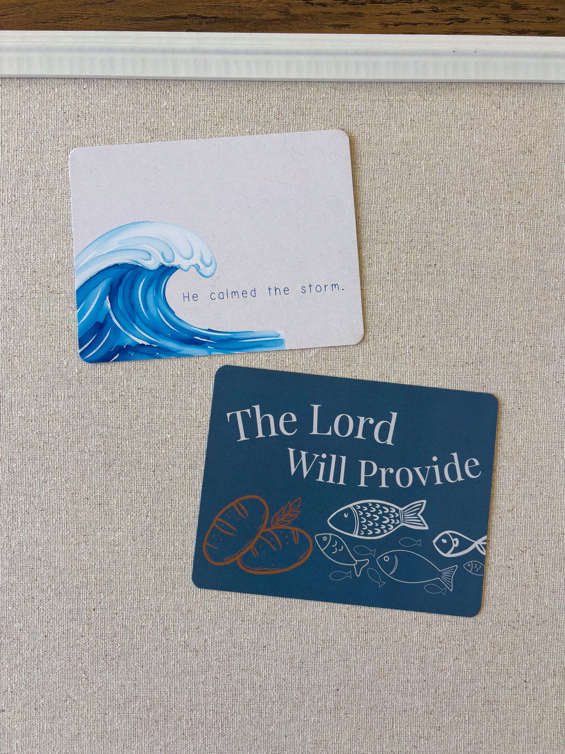 Two small printed cards or labels placed on a beige fabric surface. The top card is white with a blue ocean wave illustration and the text 'He calmed the storm.