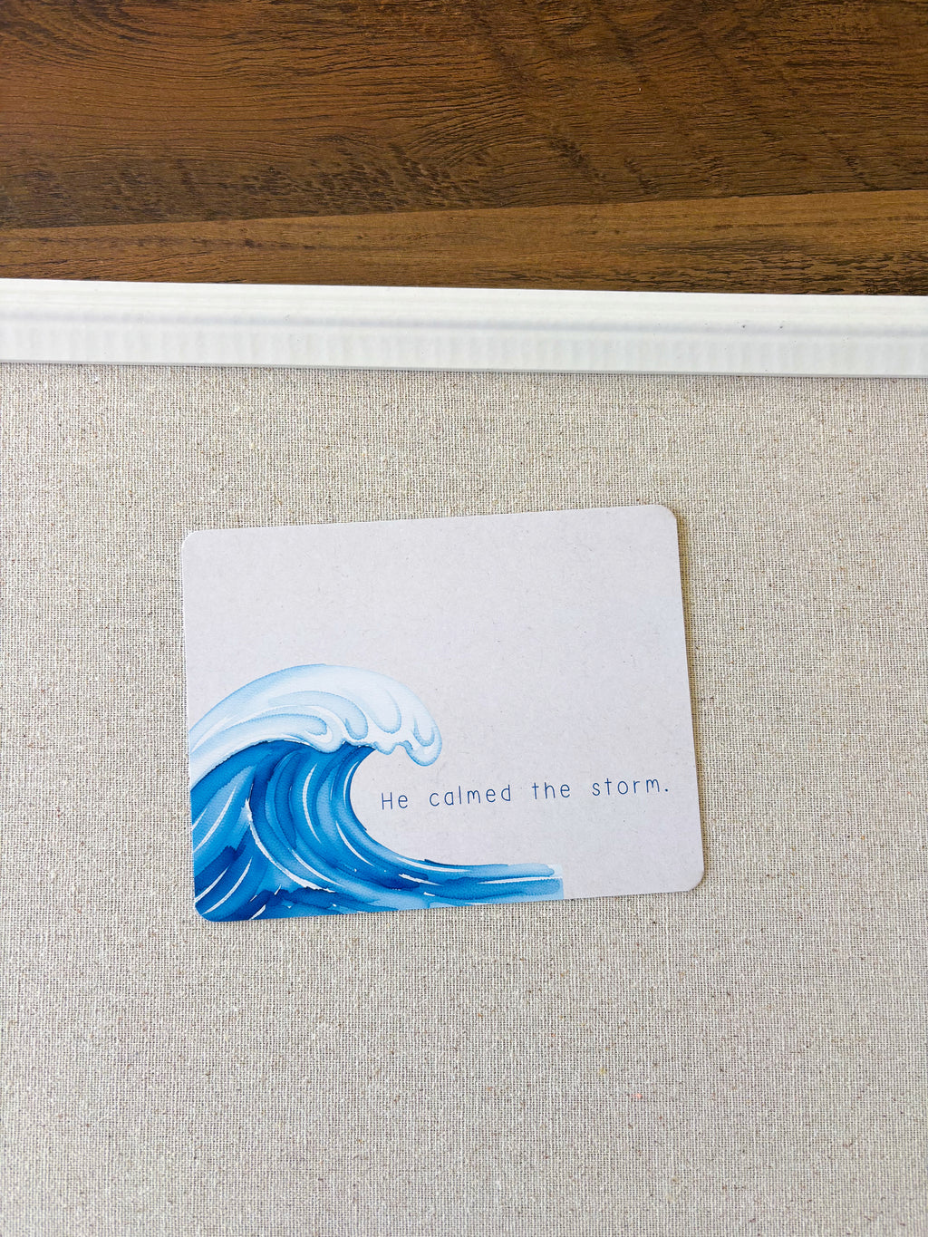 A square white card featuring a blue ocean wave illustration and the printed phrase 'He calmed the storm.' placed on a light beige textured surface with a wooden surface visible at the top.
