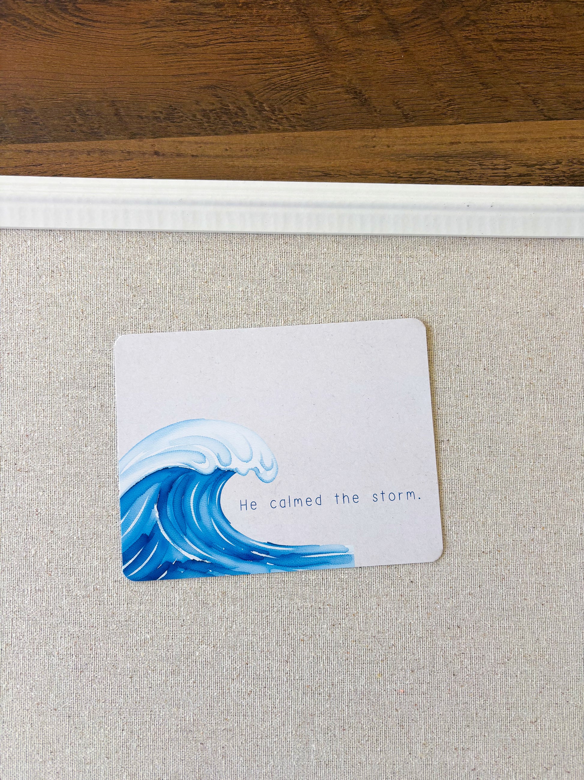 A square white card featuring a blue ocean wave illustration and the printed phrase 'He calmed the storm.' placed on a light beige textured surface with a wooden surface visible at the top.