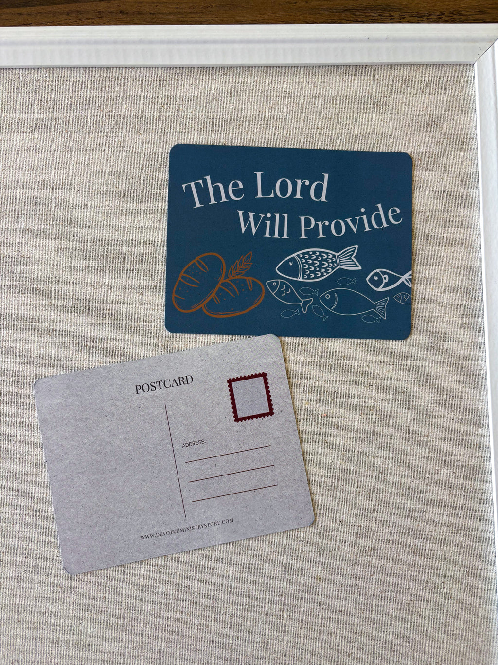 A framed display featuring a dark blue postcard with the words 'The Lord Will Provide' and simple illustrations of bread and fish, alongside a gray postcard with a red stamp box, mounted on a beige textured board.