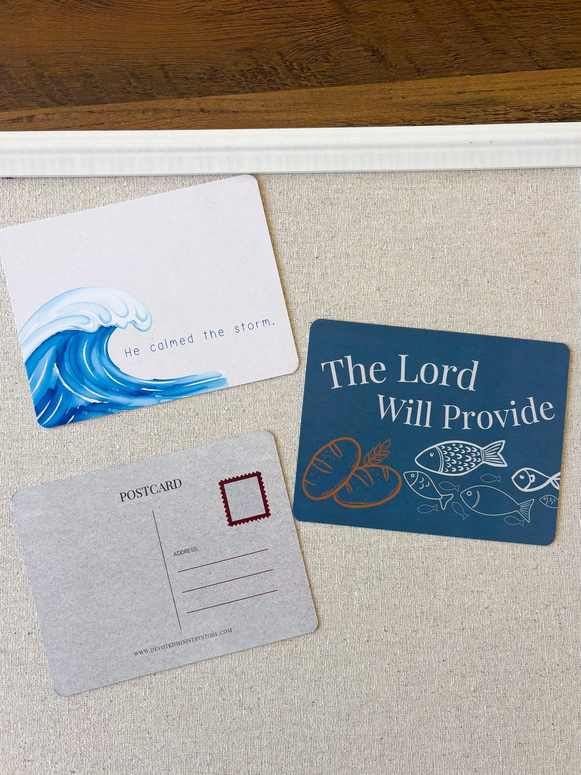 Three printed cards arranged on a light fabric surface: a white card with a blue ocean wave and the phrase 'He calmed the storm,' a gray postcard-style card with a red border box, and a dark blue card with white text reading 'The Lord Will Provide' and fish and bread illustrations.