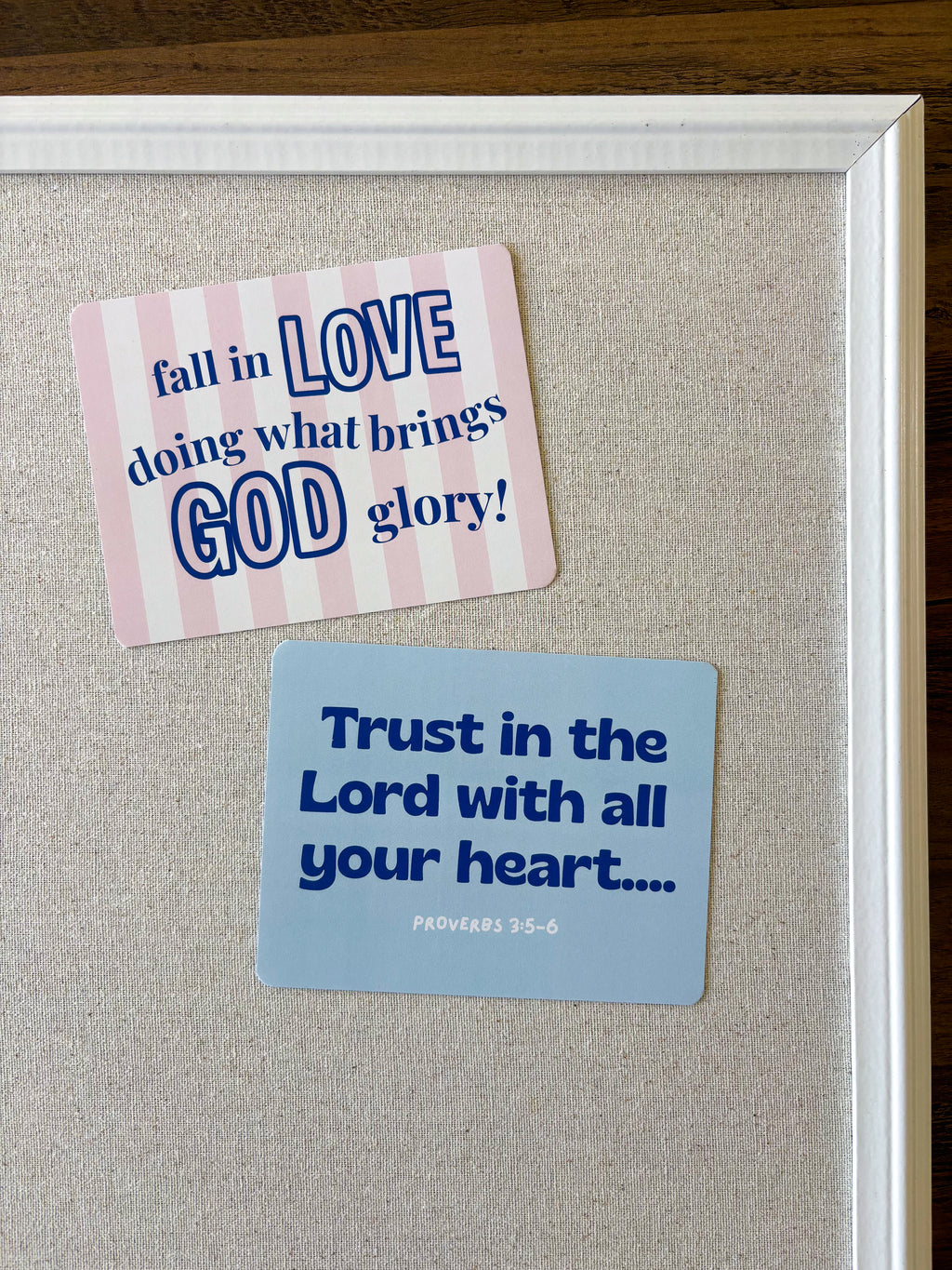 Two inspirational religious stickers displayed on a beige linen board inside a white frame. The top sticker has pink and white stripes with blue text reading 'fall in LOVE doing what brings GOD glory!'.