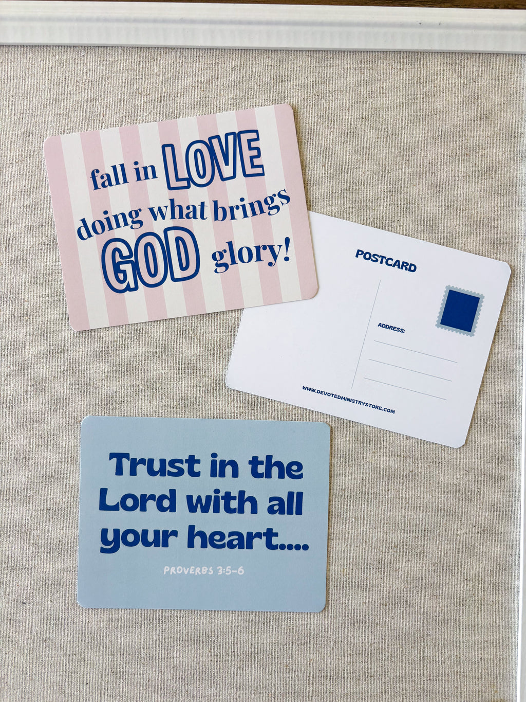 Two printed postcards displayed on a beige fabric surface. One postcard has a pink and white striped background with blue text reading 'fall in LOVE doing what brings GOD glory!'.