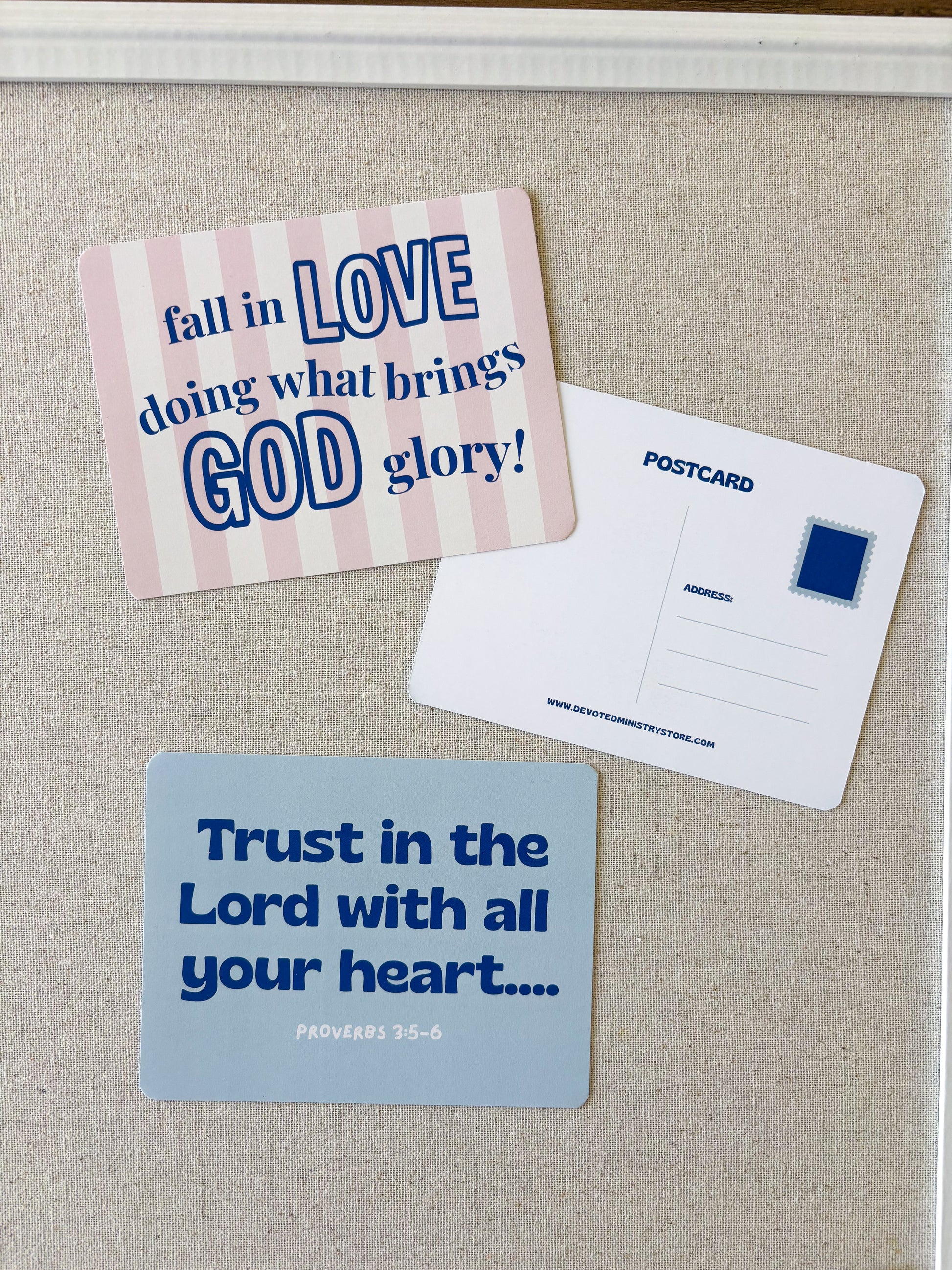 Two printed postcards displayed on a beige fabric surface. One postcard has a pink and white striped background with blue text reading 'fall in LOVE doing what brings GOD glory!'.