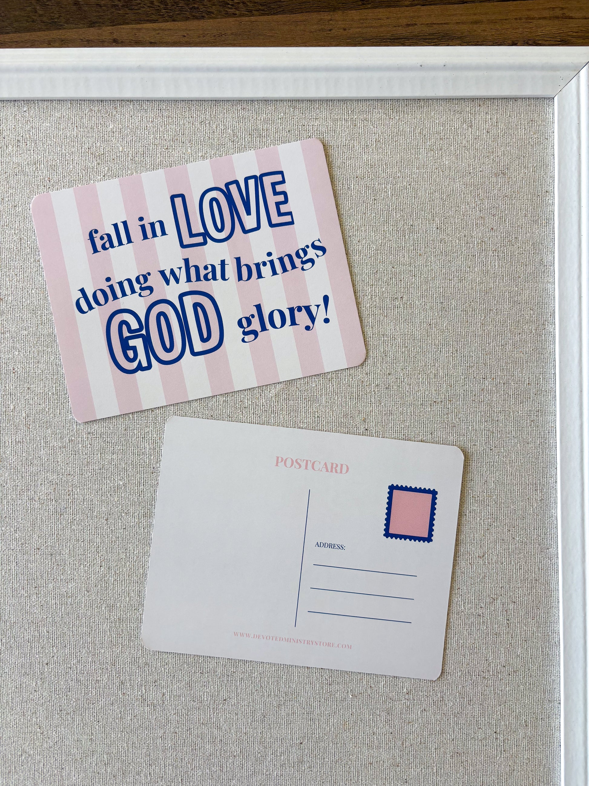A pink and white striped postcard with the text 'fall in LOVE doing what brings GOD glory!' displayed on a beige fabric-covered board next to a blank postcard with a pink stamp box.