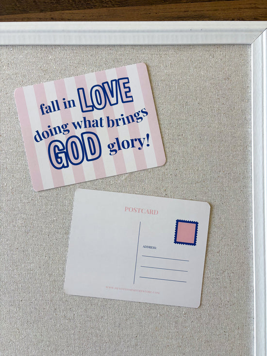A pink and white striped postcard with the text 'fall in LOVE doing what brings GOD glory!' displayed on a beige fabric-covered board next to a blank postcard with a pink stamp box.