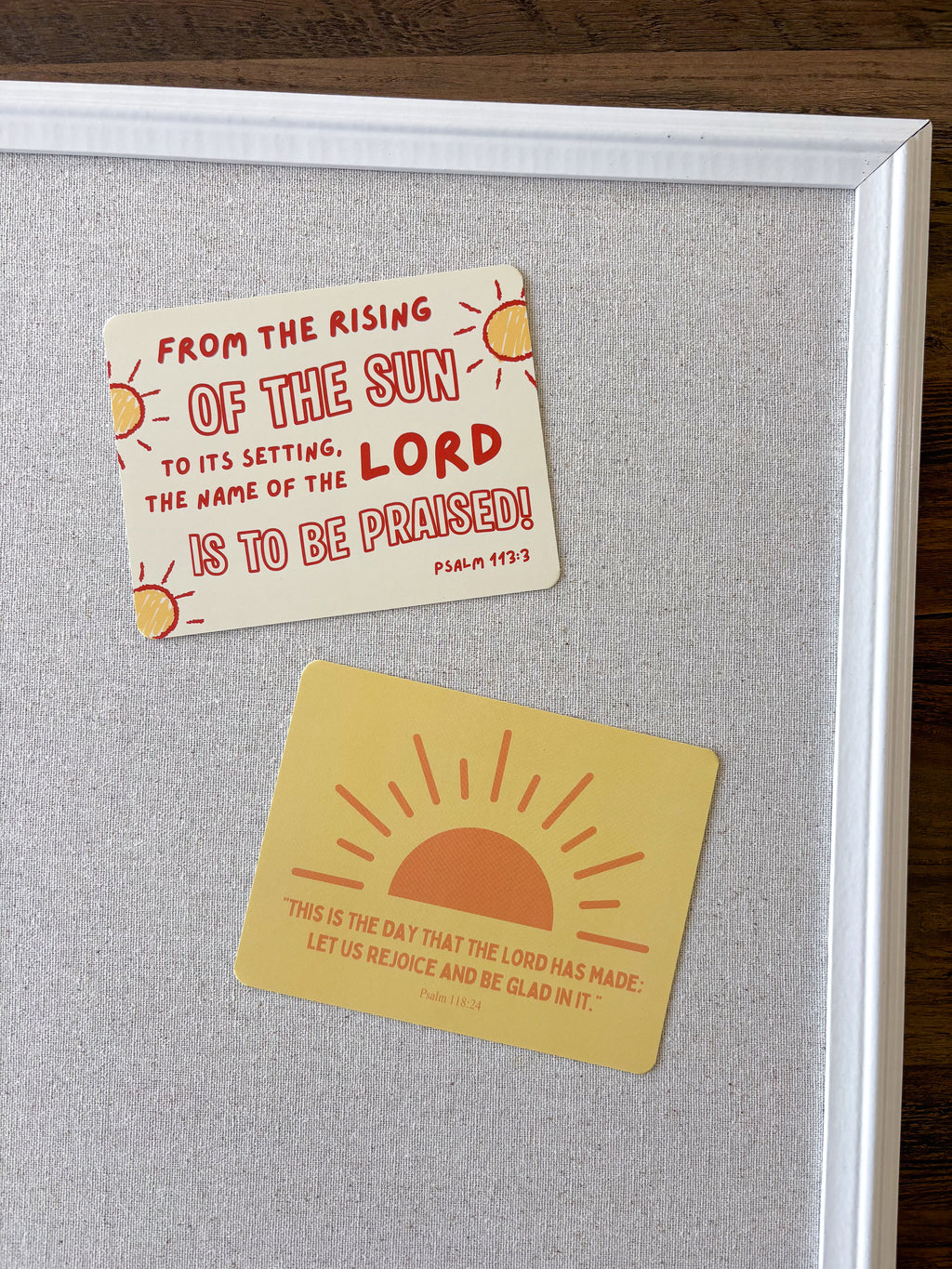 Two rectangular inspirational stickers with sun graphics and Bible verses are displayed on a gray cork board inside a white frame, set on a dark wood surface.