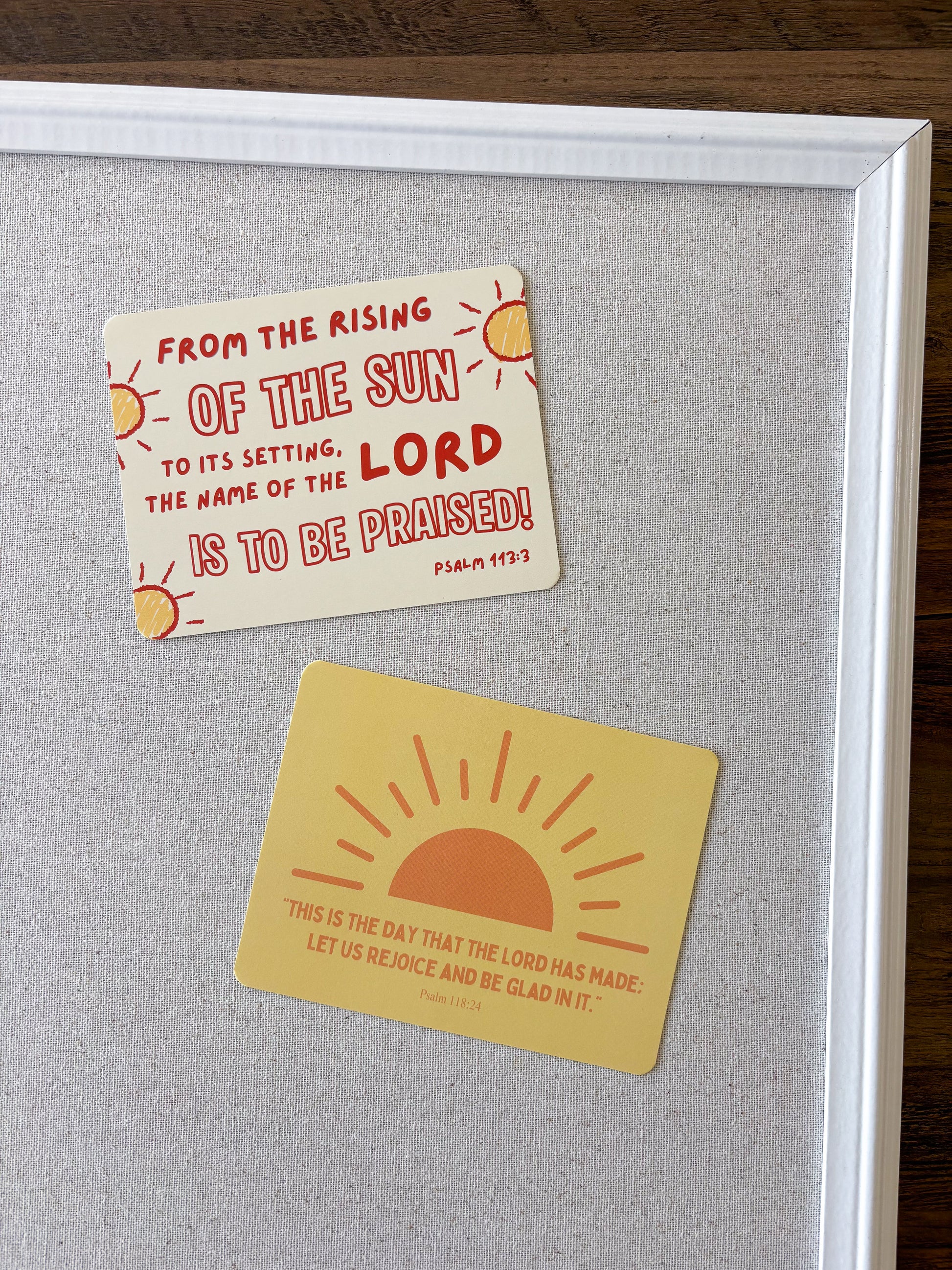 Two rectangular inspirational stickers with sun graphics and Bible verses are displayed on a gray cork board inside a white frame, set on a dark wood surface.
