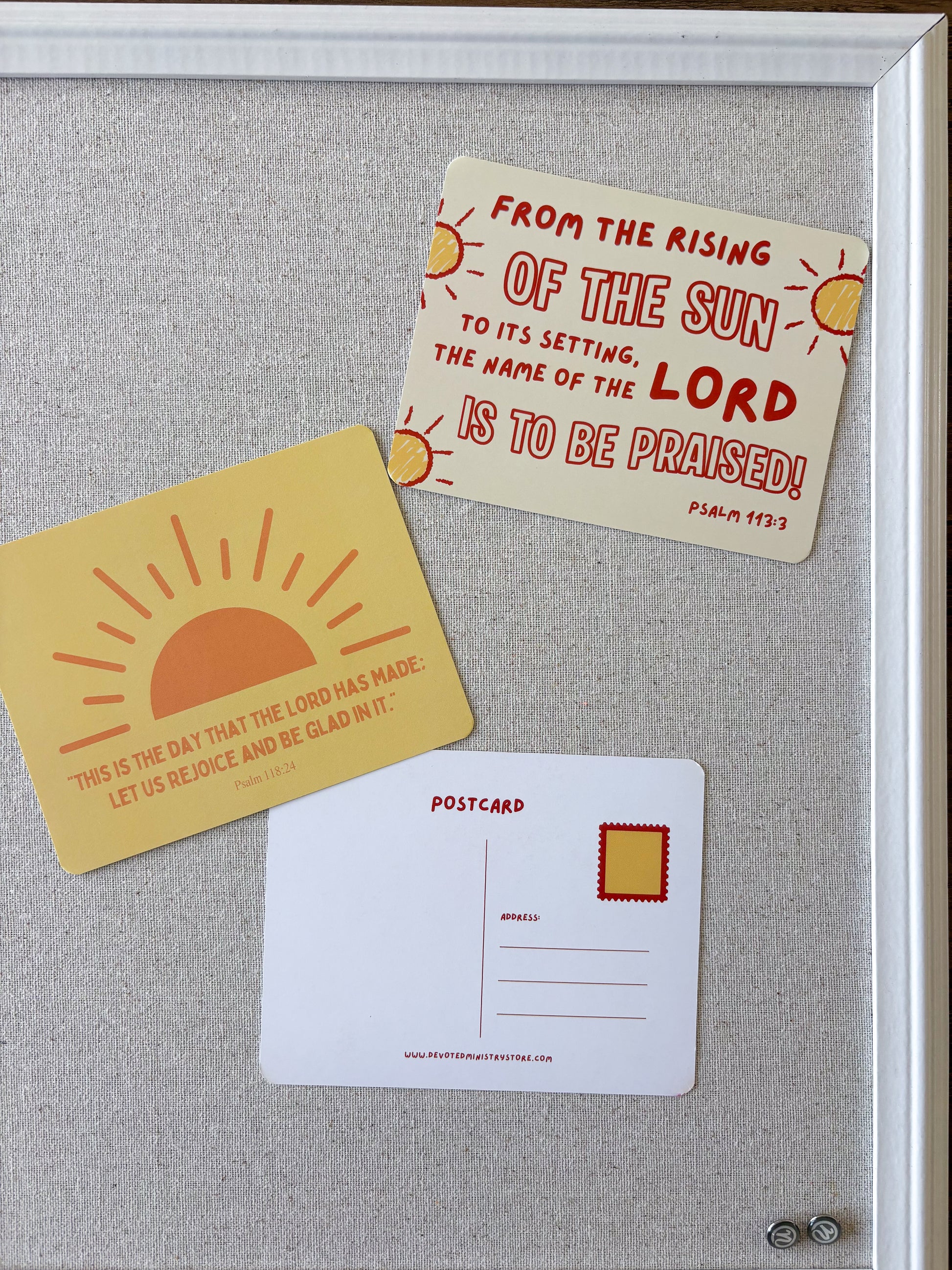 Three decorative postcards featuring Bible verses are arranged on a light gray fabric surface inside a white frame. One postcard is pale yellow with a red sun illustration and a scripture quote from Psalm 103:30.
