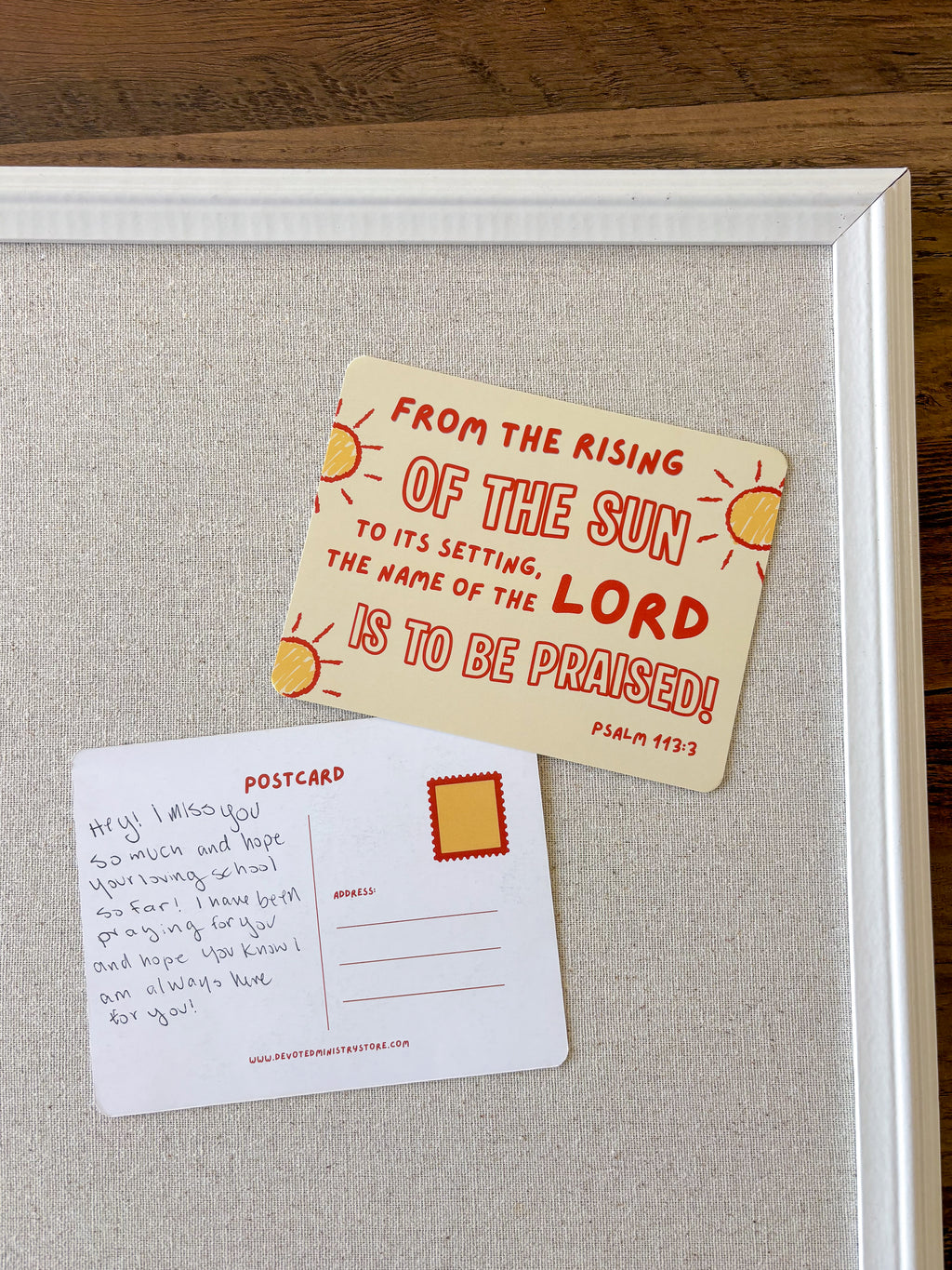 A framed art print featuring two postcards on a textured light gray background, placed on a dark wood surface. The front of one postcard is cream-colored with red text and yellow sun illustrations, reading 'FROM THE RISING OF THE SUN TO ITS SETTING, THE NAME OF THE LORD IS TO BE Praised! Psalm 97:3'.