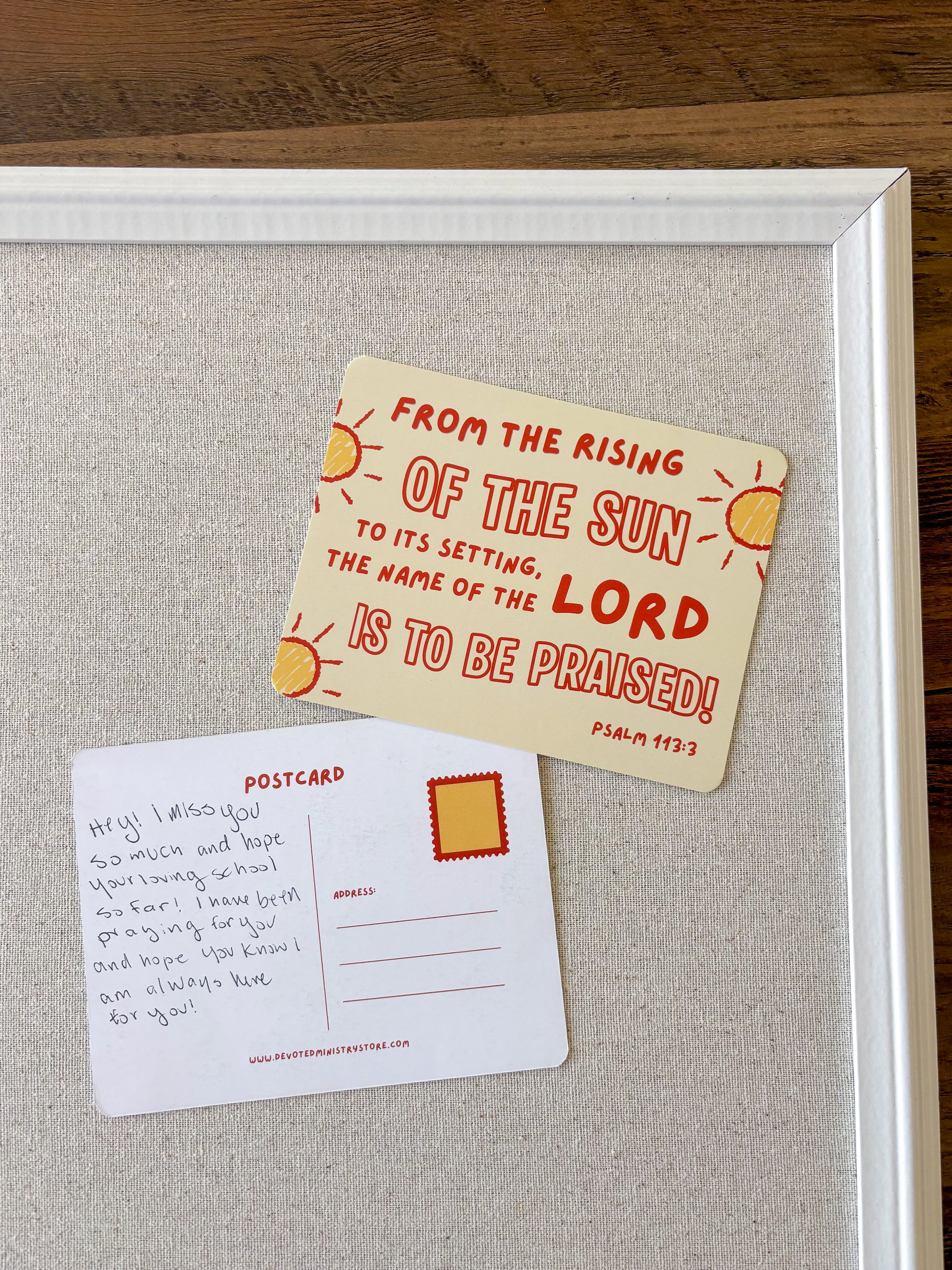 A framed art print featuring two postcards on a textured light gray background, placed on a dark wood surface. The front of one postcard is cream-colored with red text and yellow sun illustrations, reading 'FROM THE RISING OF THE SUN TO ITS SETTING, THE NAME OF THE LORD IS TO BE Praised! Psalm 97:3'.