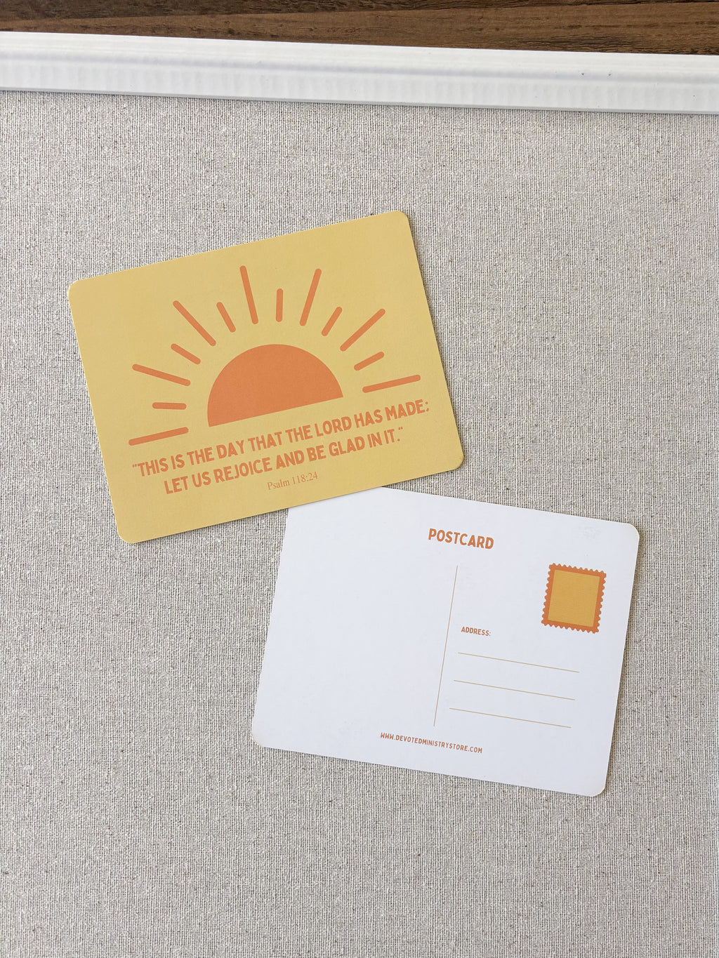 Two postcards displayed on a textured light gray fabric surface. The front card is pale yellow with an orange sun graphic and a Bible verse from Psalm 148:13.