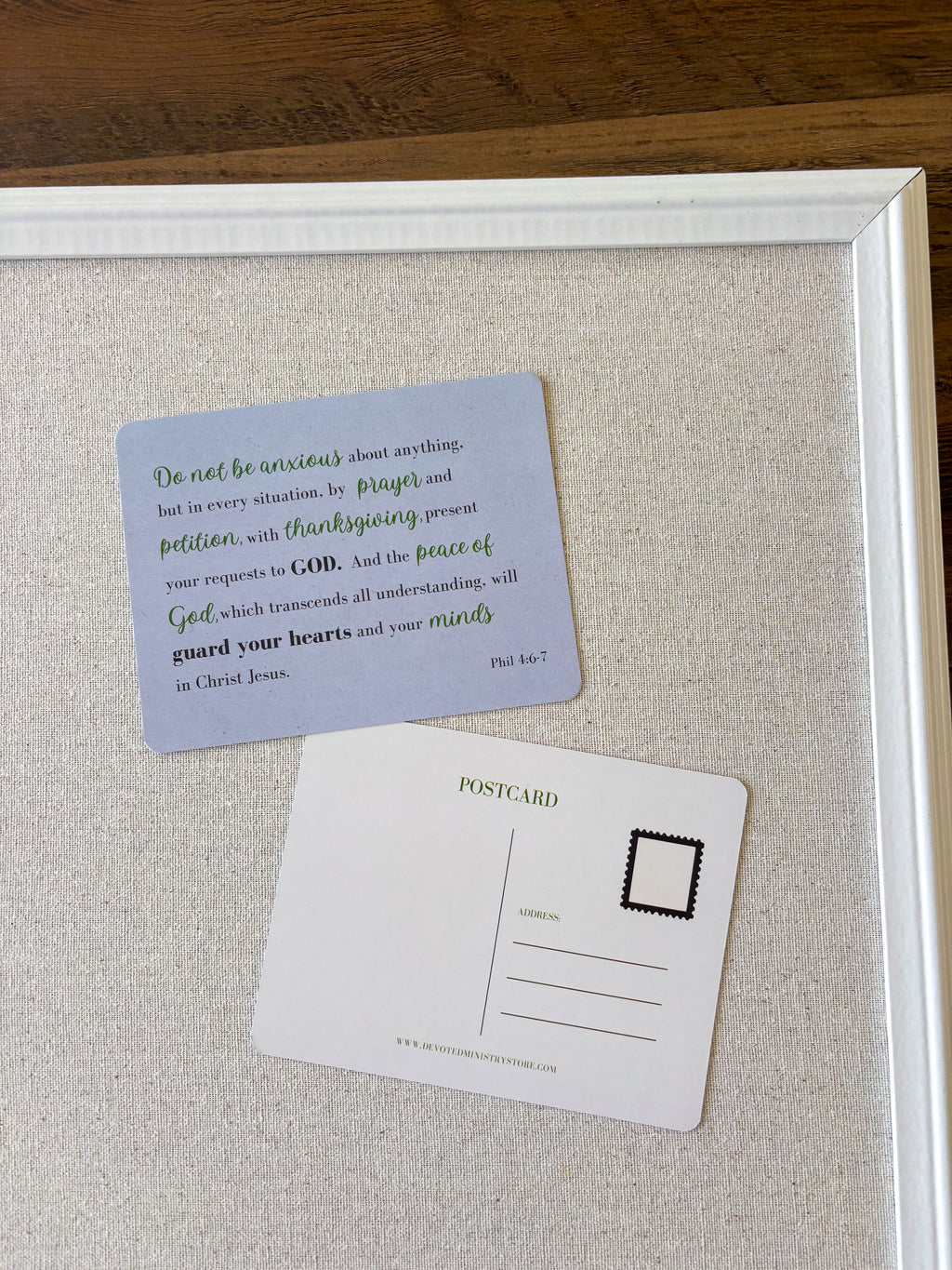A framed art print featuring a light blue postcard with a Bible verse and a white postcard on a textured beige background, all enclosed in a white frame.