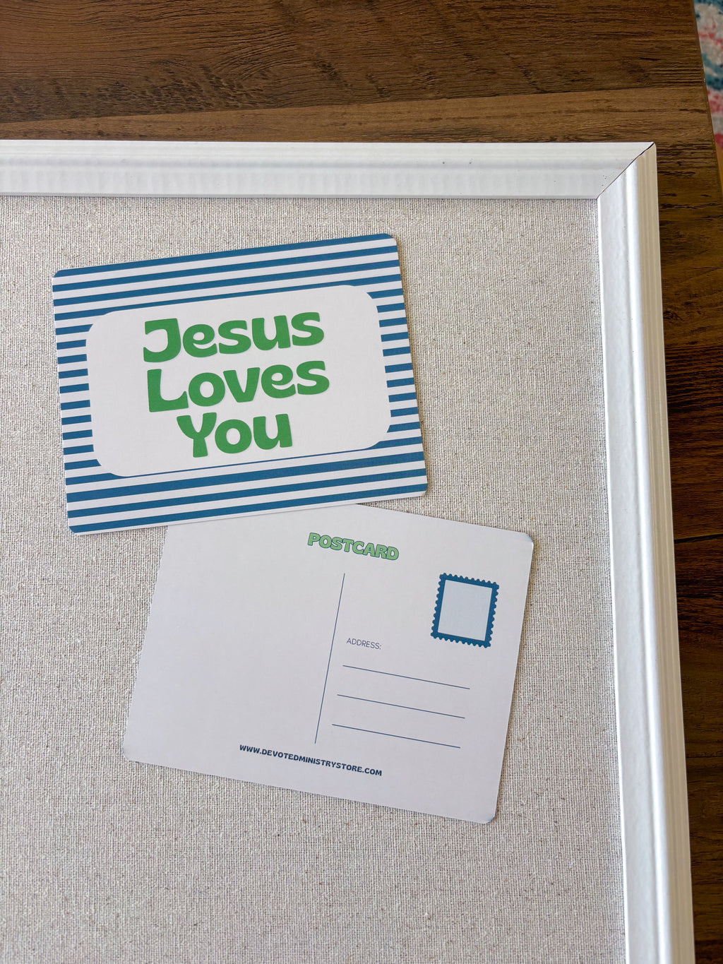 A framed display with two postcard-style signs on a beige fabric-covered board. One card is blue and white striped with the words 'Jesus Loves You' in green, and the other is a blank white postcard with a blue stamp box and green 'POSTCARD' text.