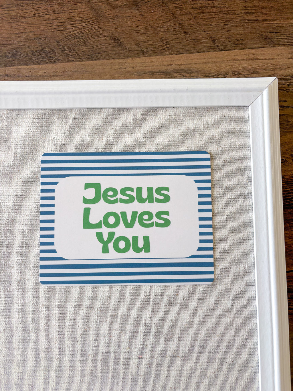 A square decorative sign with the words 'Jesus Loves You' in green text on a white rounded rectangle, bordered by blue horizontal stripes. The sign is mounted on a light beige textured board inside a white frame, set against a dark wood background.