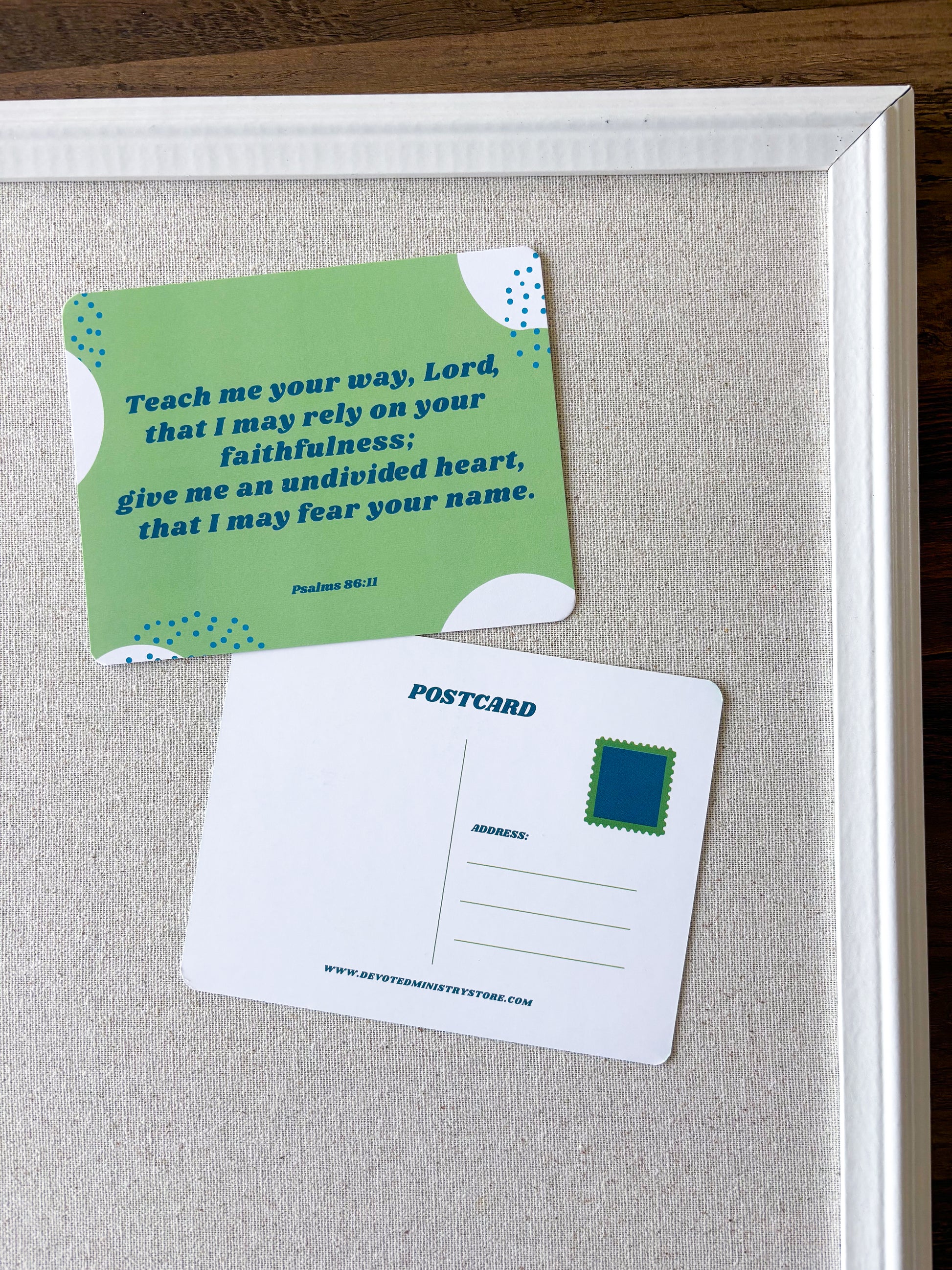 A framed decorative display featuring a green postcard-style card with a Bible verse and a white postcard placed on a light beige fabric background inside a white frame.