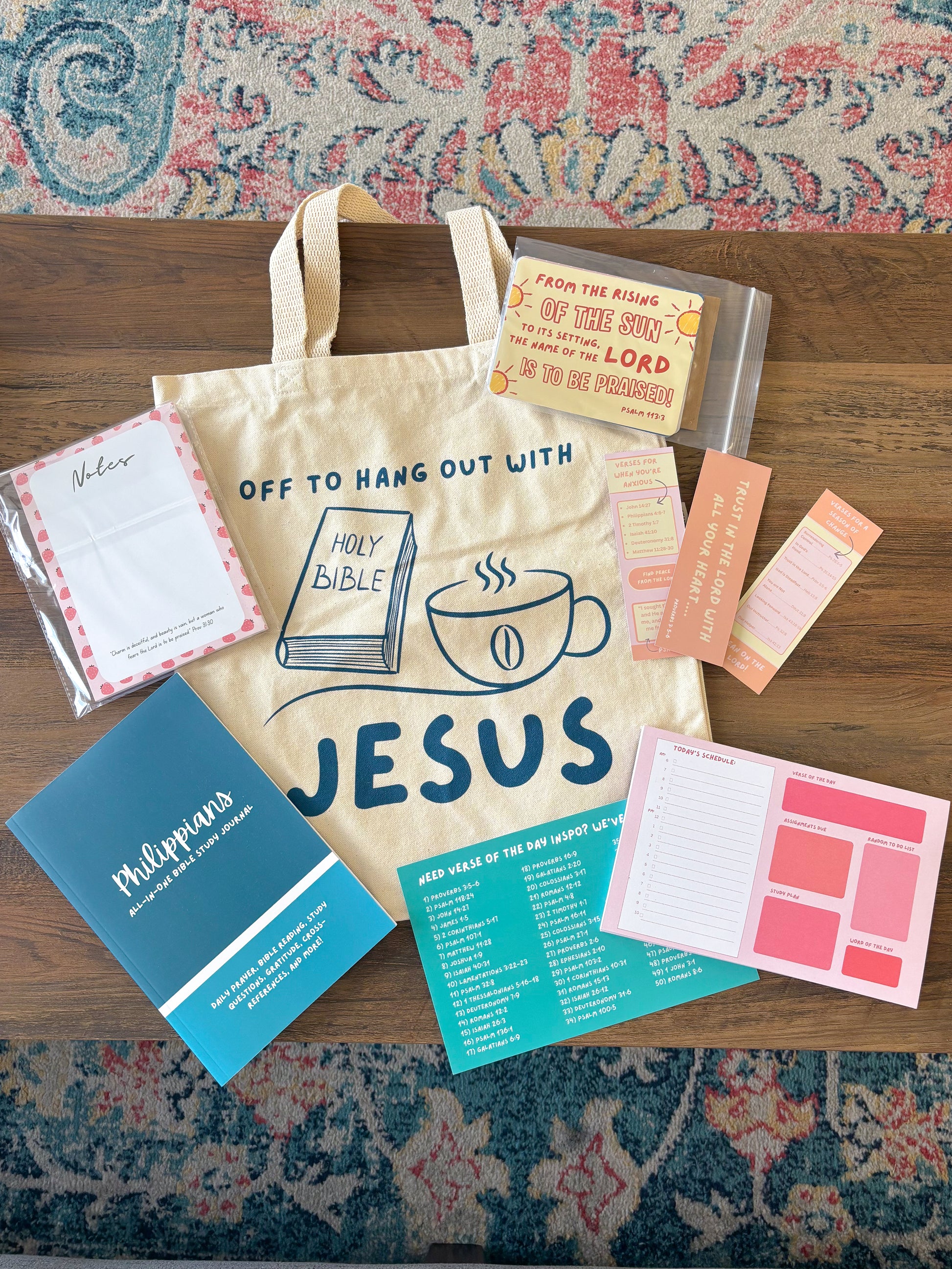 A flat-lay display of a personalized canvas tote bag with religious-themed stationery and Bible study materials arranged on a wooden table over a patterned rug. The tote bag is cream-colored with navy blue text and graphics reading 'OFF TO HANG OUT WITH JESUS' and features a drawing of a Holy Bible and coffee cup.