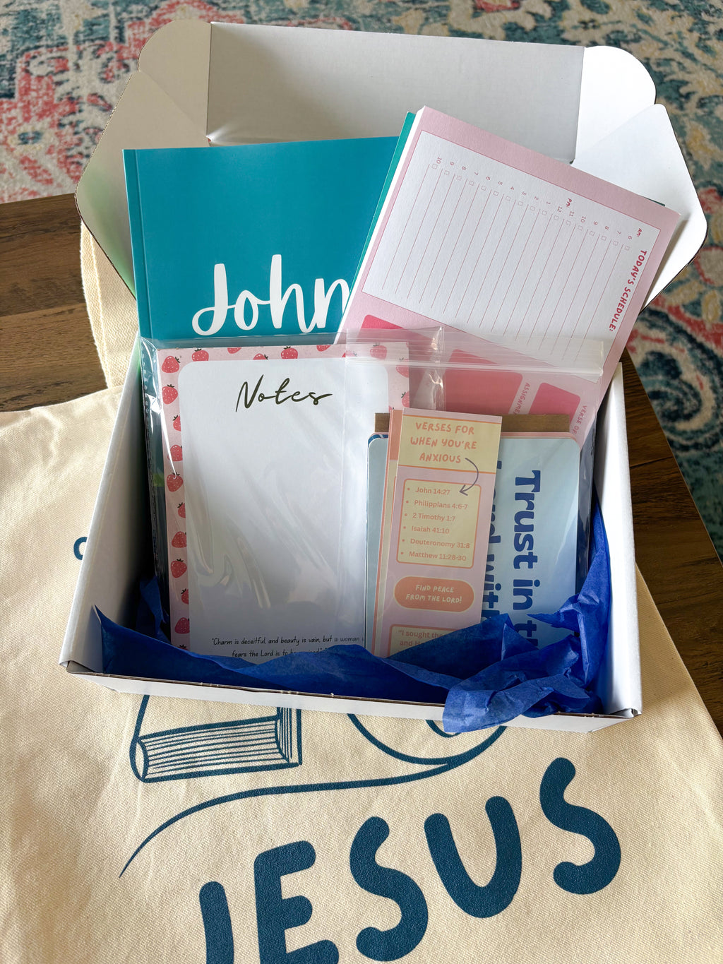 A white cardboard gift box filled with stationery items, including a teal notepad, pink lined notes, sticky notes, and a planner insert, set on a beige tote bag with the word 'JESUS' printed on it.