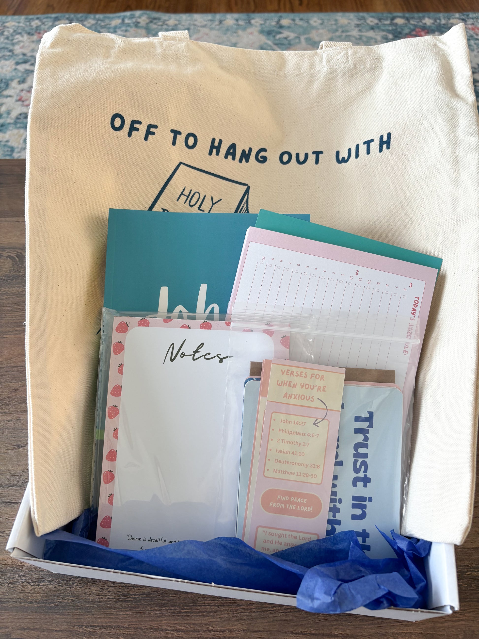 A cream canvas tote bag printed with 'OFF TO HANG OUT WITH Jesus' sits inside a white gift box filled with stationery items, including a teal card, lined notes, a pink memo pad, and blue tissue paper.
