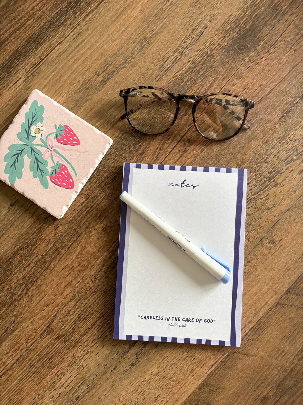 blue stripped notepad with a Bible verse on a brown desk with glasses and a fruit coaster 