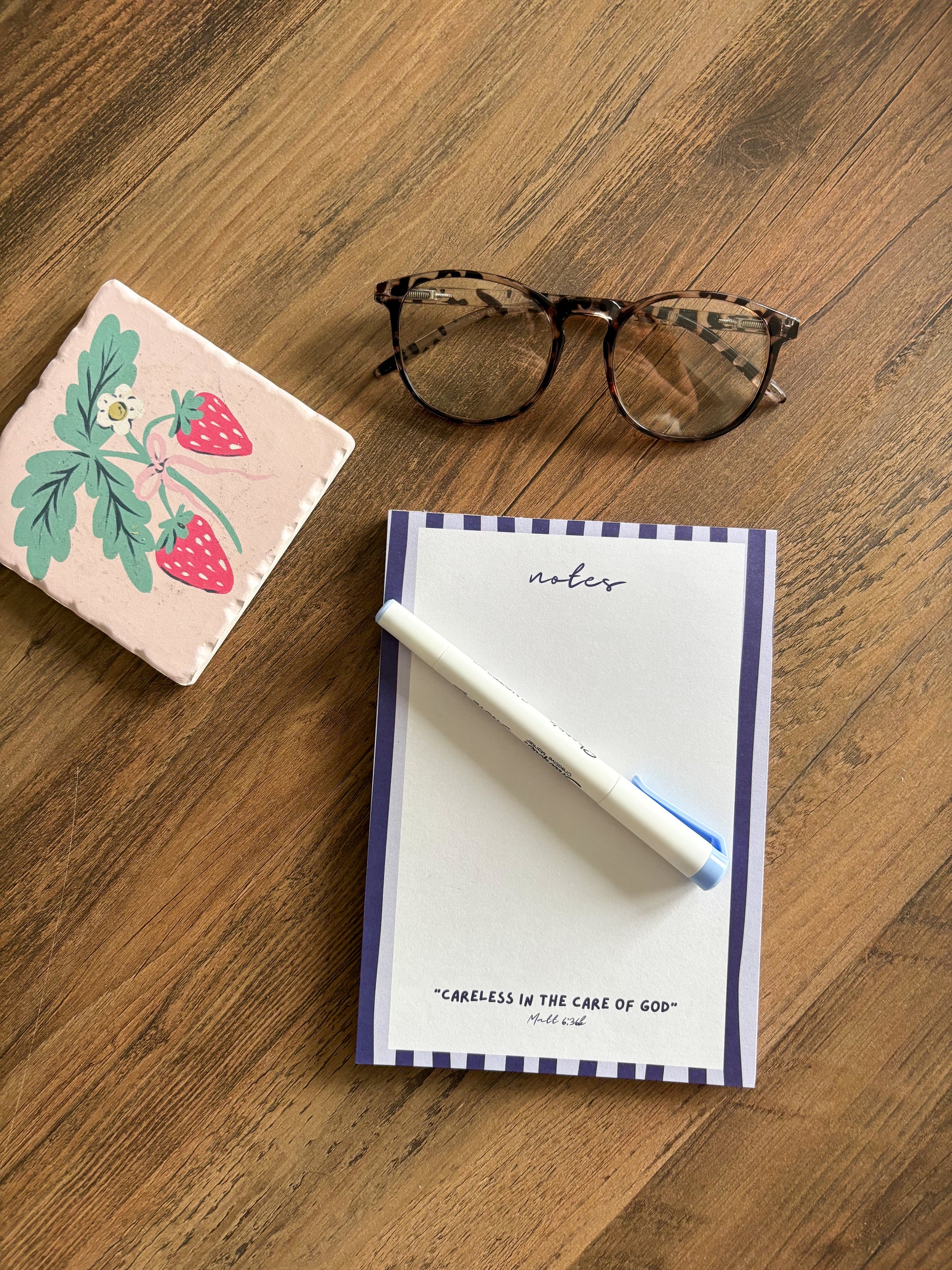 blue stripped notepad with a Bible verse on a brown desk with glasses and a fruit coaster 