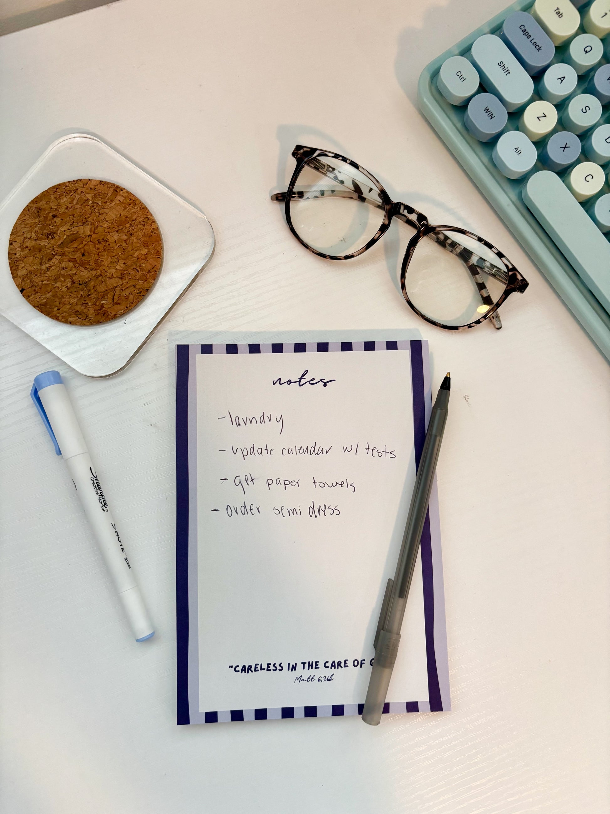 blue striped notepad with a Bible verse and pen on a white desk with glasses, a keyboard and coaster on it